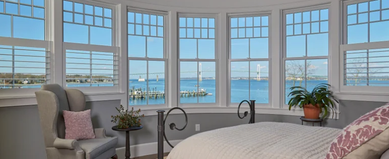 A bedroom with large windows overlooking a body of water and a bridge in the distance, featuring a bed, an armchair, and potted plants.
