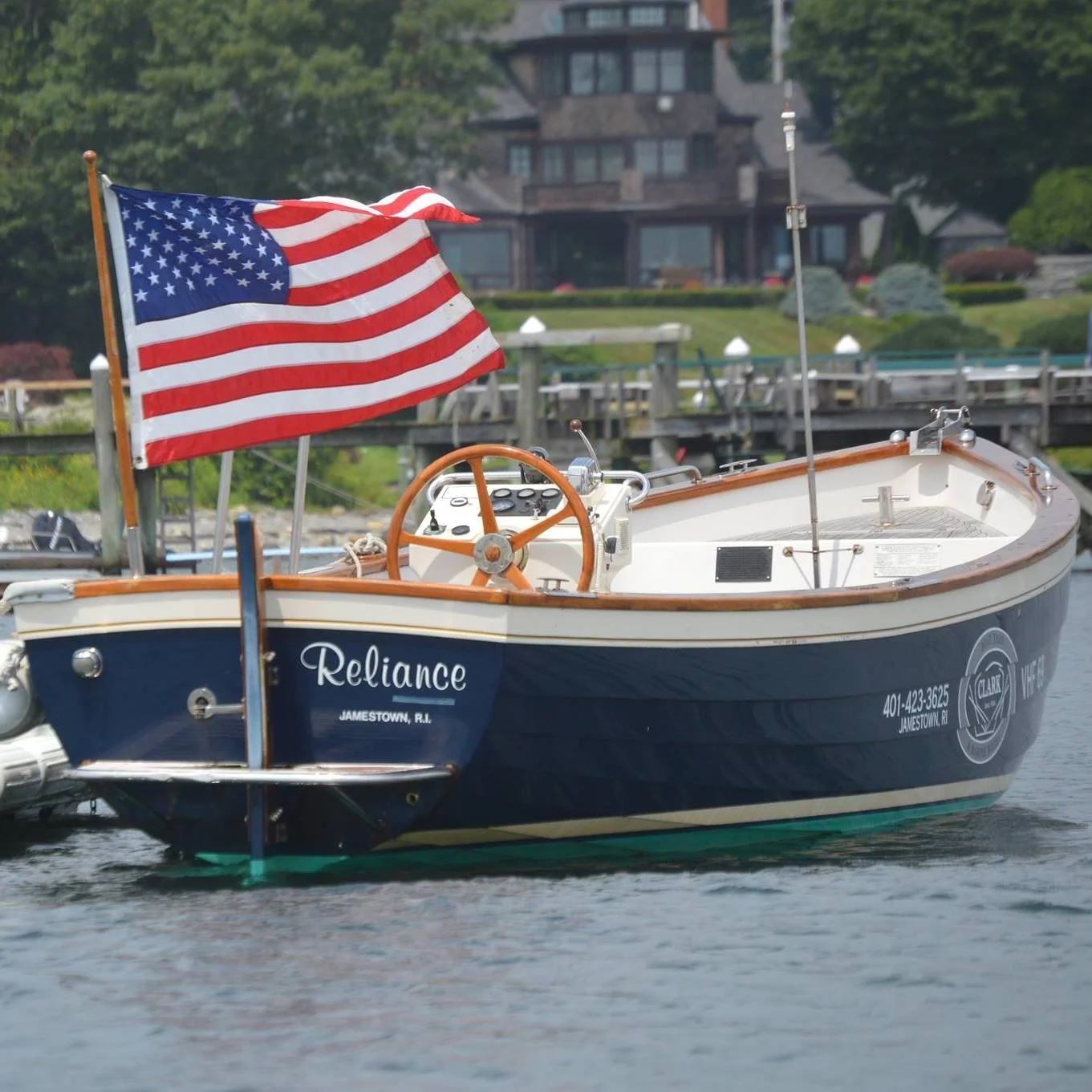 A blue and white boat named Reliance with a large American flag at the stern, floating on a body of water with a house and green trees in the background.