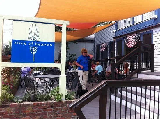 Outdoor seating area at a restaurant with a sign that reads 'slice of heaven.' There are several tables and chairs, with some people sitting and standing around. The area is shaded with orange and yellow fabric canopies, and there's a staircase leading to the restaurant. The setting appears to be in a small town or neighborhood.