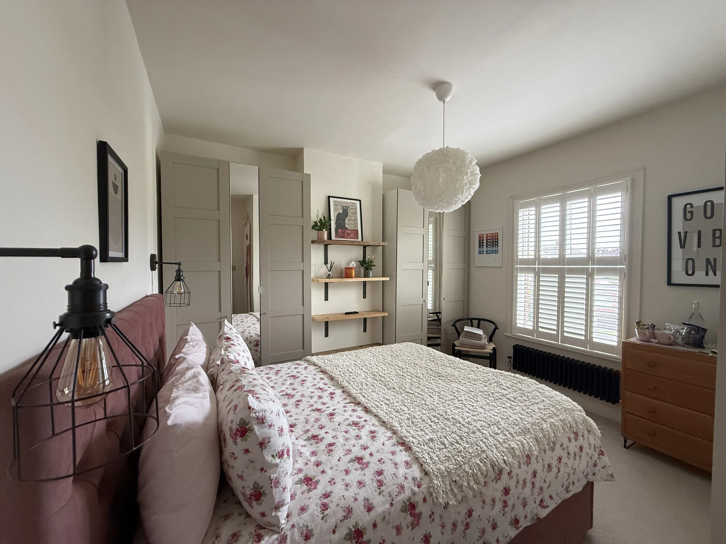A bedroom with a large bed featuring a floral quilt and pillows, a pink headboard, black wire wall-mounted lamps, a wooden dresser, and large windows with white shutters. The room has neutral-colored walls, a white textured blanket on the bed, floating shelves with decorative items, and framed art, creating a cozy and modern space.