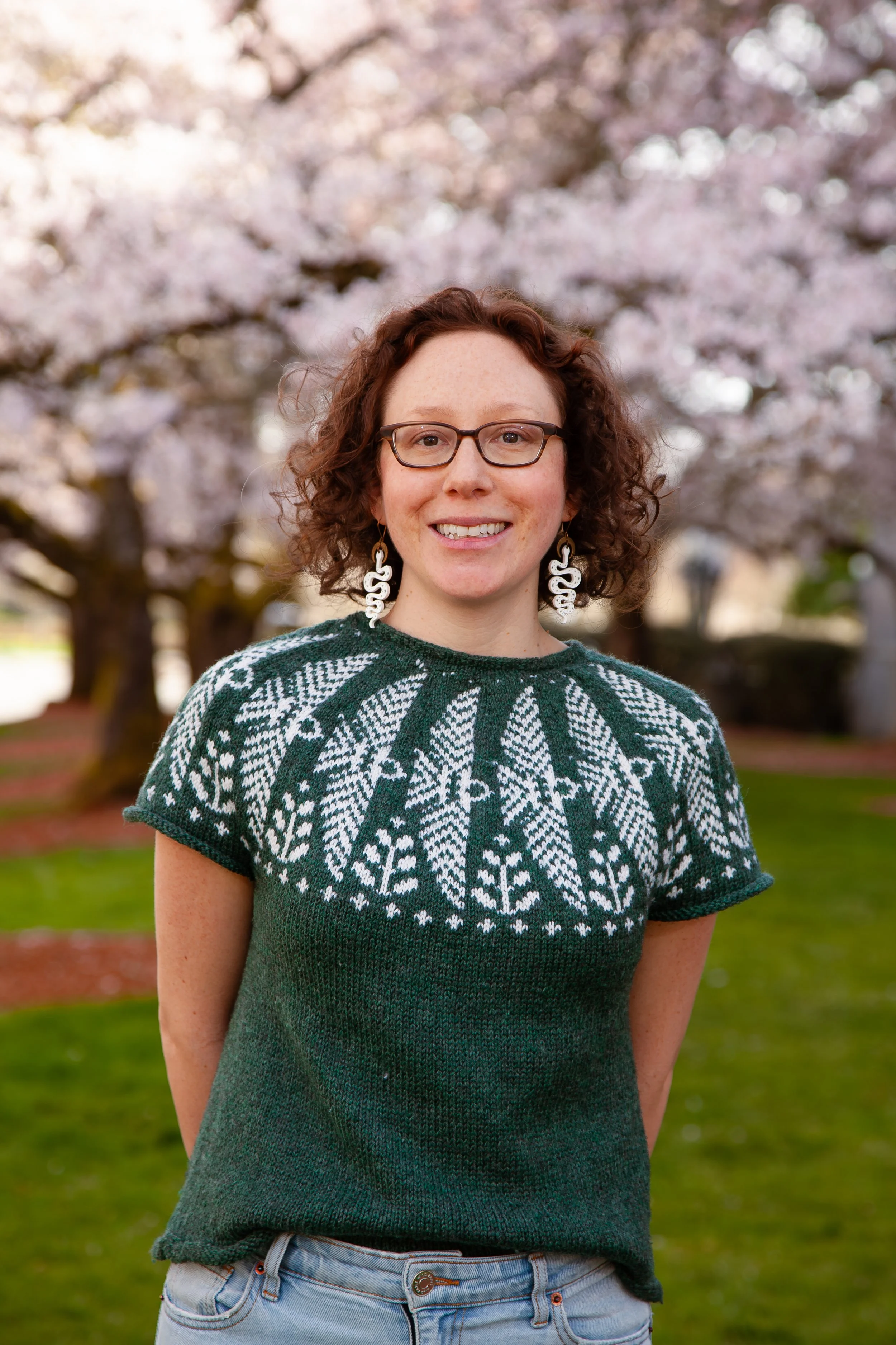 A woman with curly red hair and glasses sitting at a wooden desk, holding a book, in a cozy home office decorated with a vase of flowers, a lit candle, and a wall hanging.