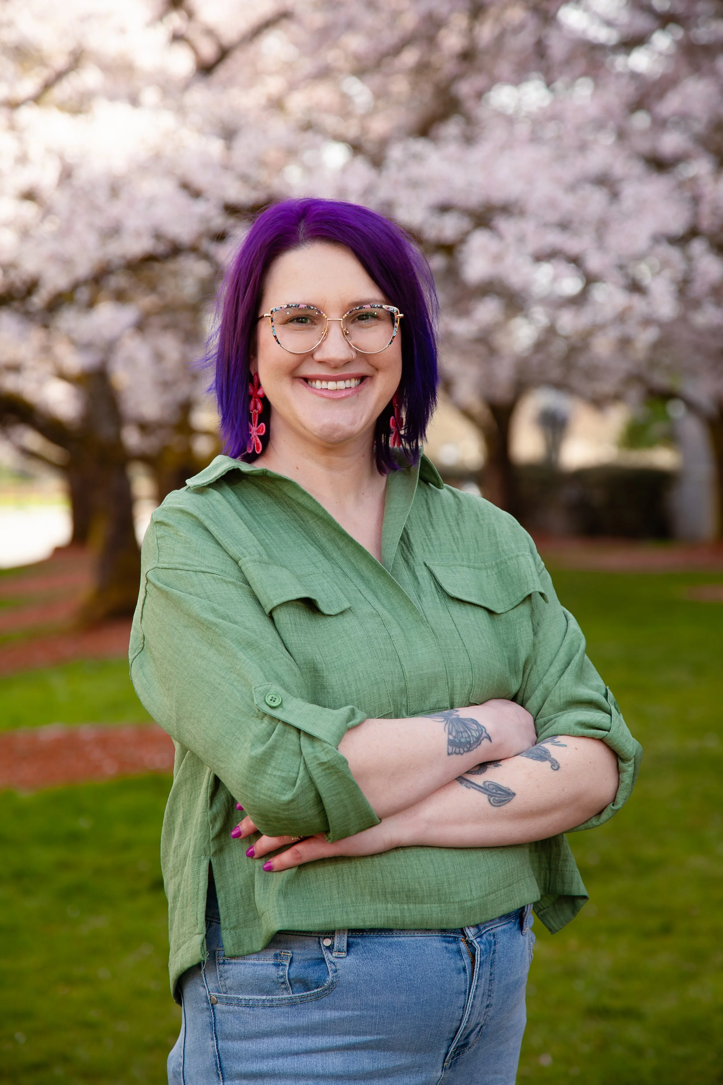 A smiling woman with purple hair, glasses, and tattoos on her arms standing outdoors in front of a garden with colorful plants during golden hour.