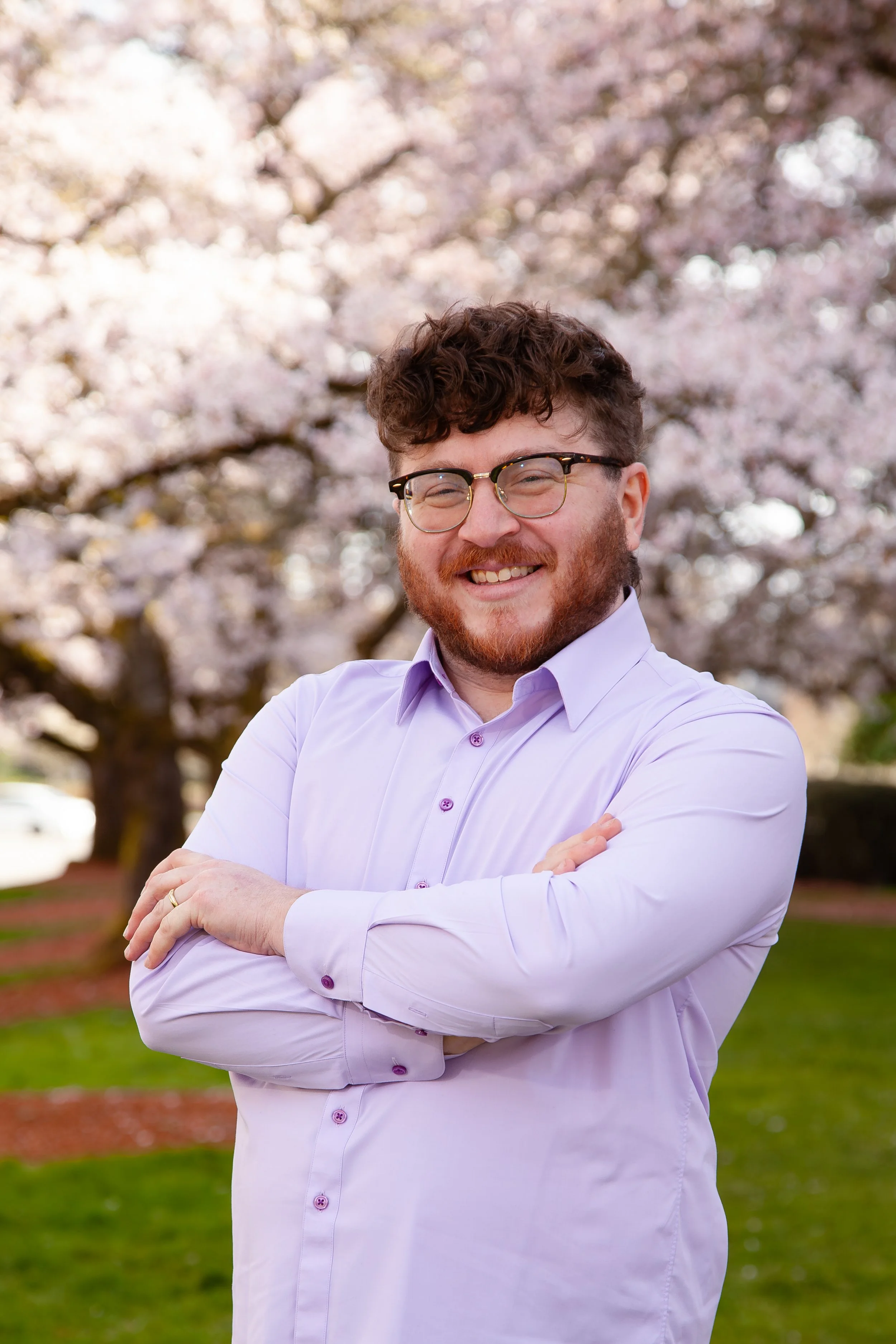 A young man with curly hair, glasses, and a beard, smiling outdoors in front of blooming flowers and greenery.