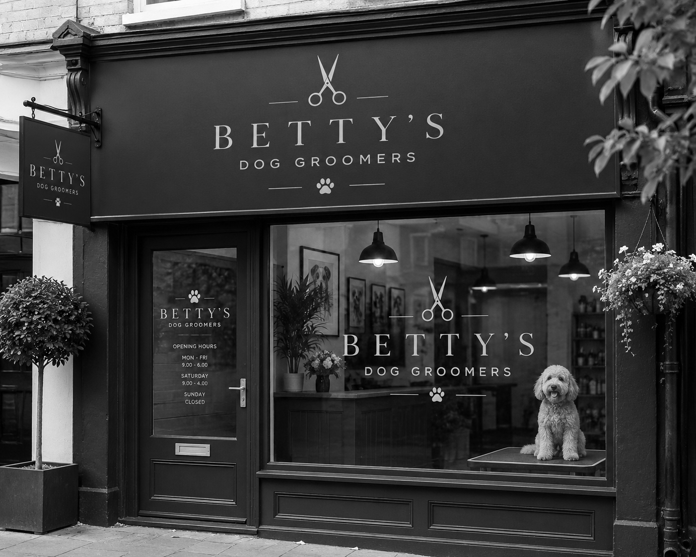 Black and white photo of a storefront for Betty's Dog Groomers, with a large window displaying a dog sitting on a grooming table. The store has hanging lights, potted plants, and framed pictures inside. The storefront signage features scissors and paw print icons.