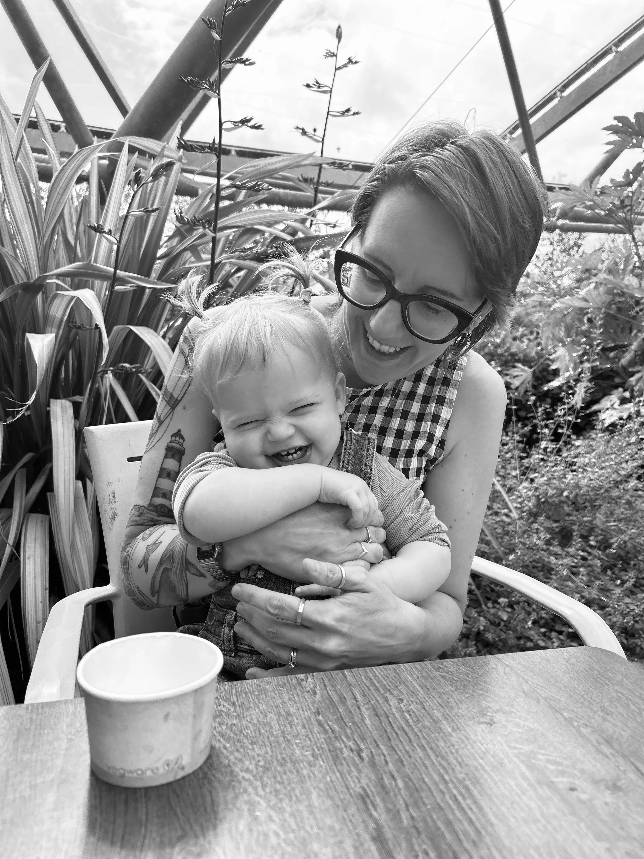 A woman with glasses and short hair holding a young, smiling child with pigtails in an outdoor or greenhouse setting with plants and a cloudy sky in the background.