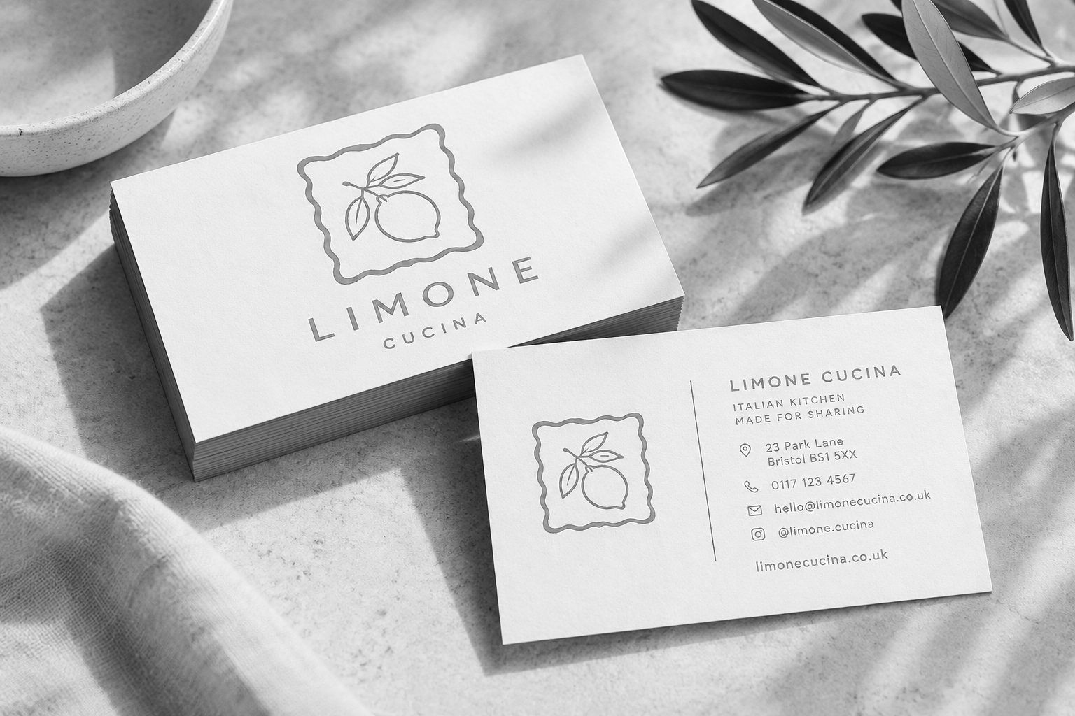 Business cards for Limone Cucina, an Italian restaurant, featuring a lemon and leaf logo. One card is placed on a textured surface, with a plant and a bowl nearby, in black and white.