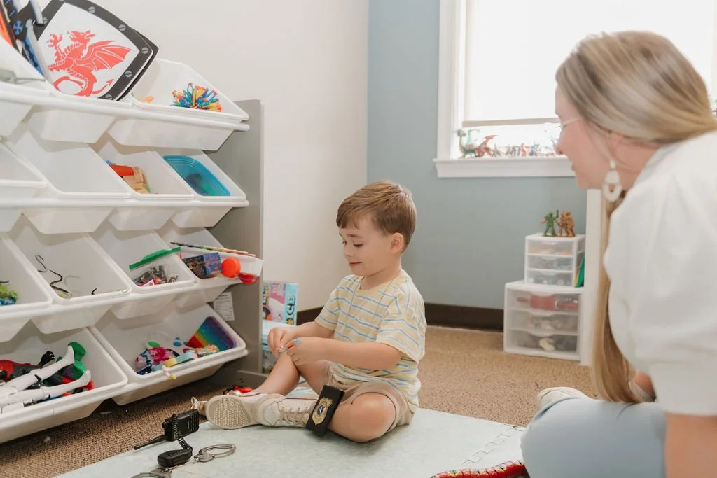 A young boy sitting on a white foam mat on the floor of a playroom, playing with toys and accessories, with a woman nearby smiling, and shelves containing toys and figurines in the background.