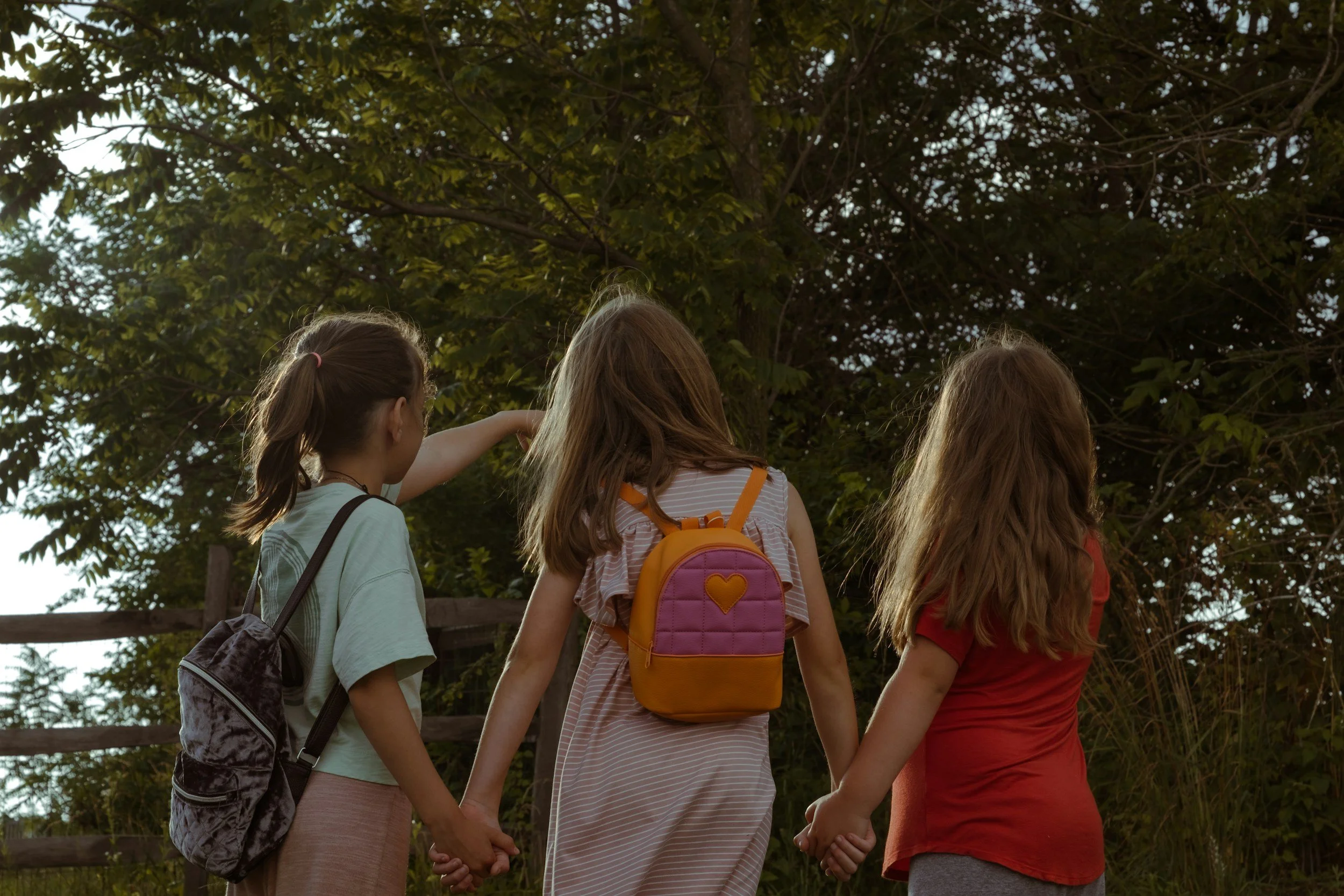 Three young girls holding hands in a circle outdoors, facing away from the camera, with trees and a wooden fence in the background.