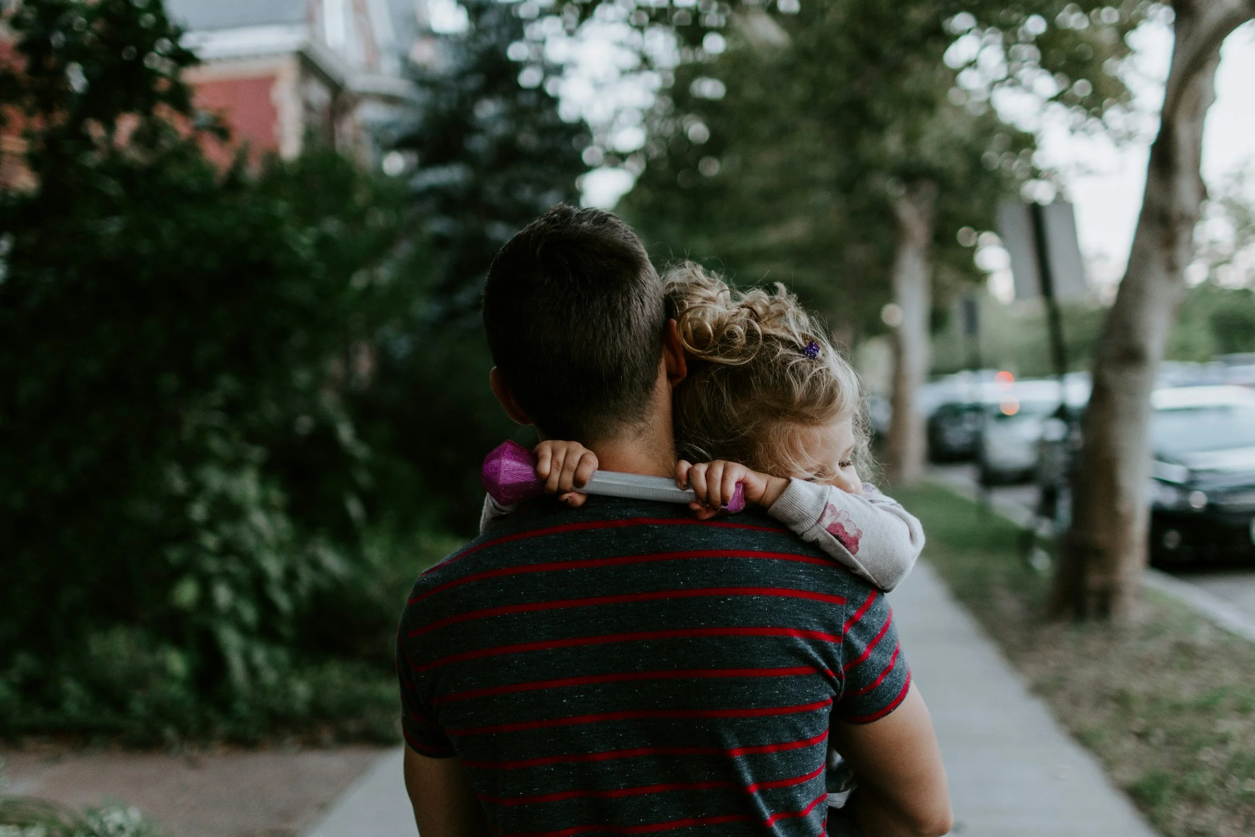 A man with short dark hair carrying a young girl with curly blonde hair and a purple bow on her head, on a sidewalk with trees and parked cars in the background.