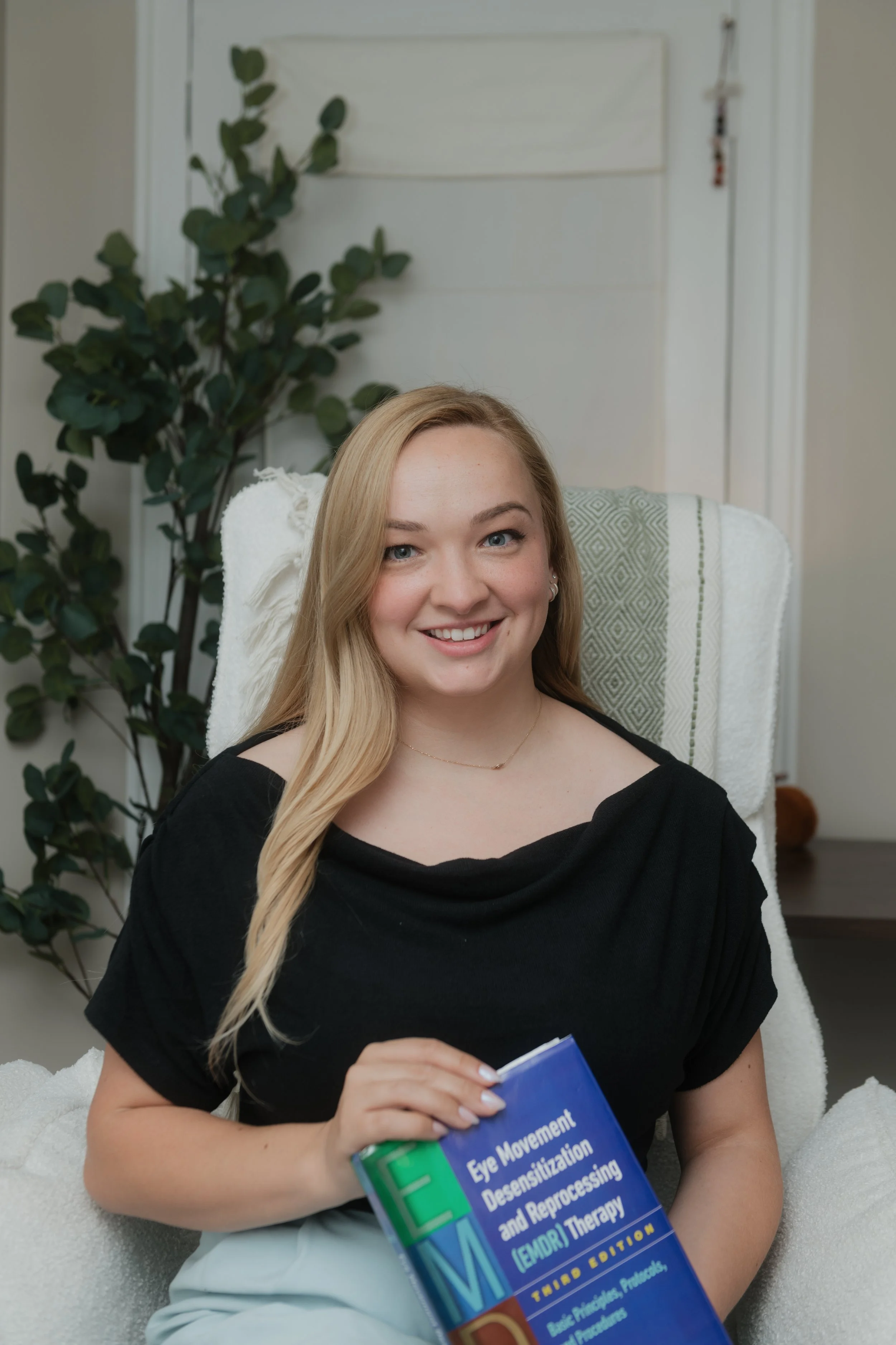 A young woman with long blonde hair, wearing a black top, seated in a white chair, holding a medical textbook titled 'Eye Movement Desensitization and Reprocessing (EMDR) Therapy'. She is smiling and looking at the camera. In the background, there is a large potted plant and a white door.