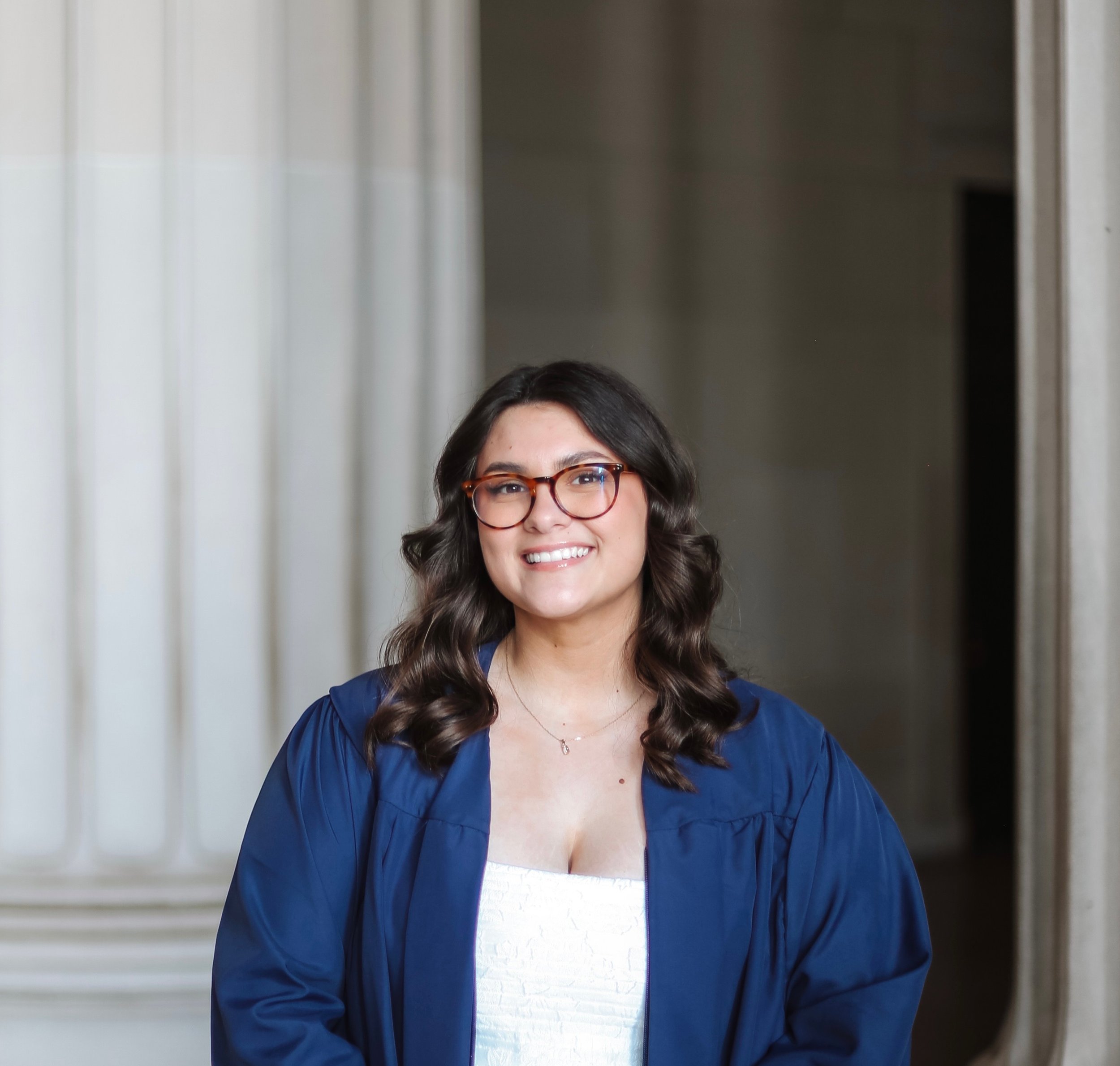 A woman with long wavy hair wearing glasses smiling, dressed in a white top and a blue jacket, standing in front of a neutral background with vertical lines.