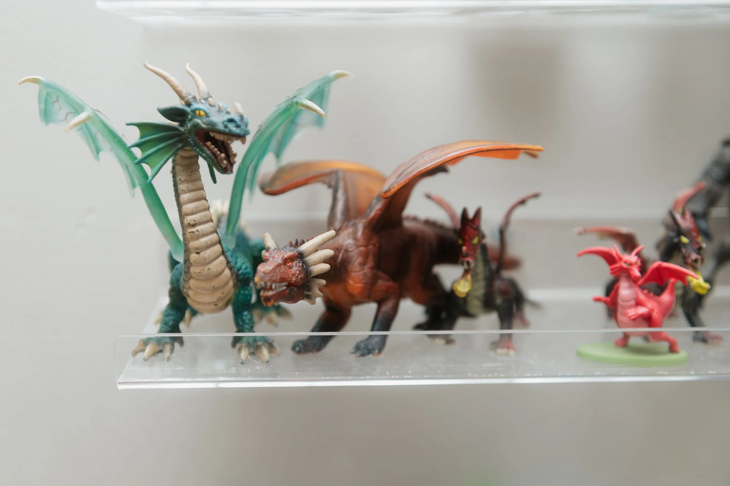 Collection of plastic or toy dinosaur figurines displayed on a clear stand, including a blue and green dragon with horns and wings, a brown dinosaur with a frill, and smaller red and black dinosaurs.