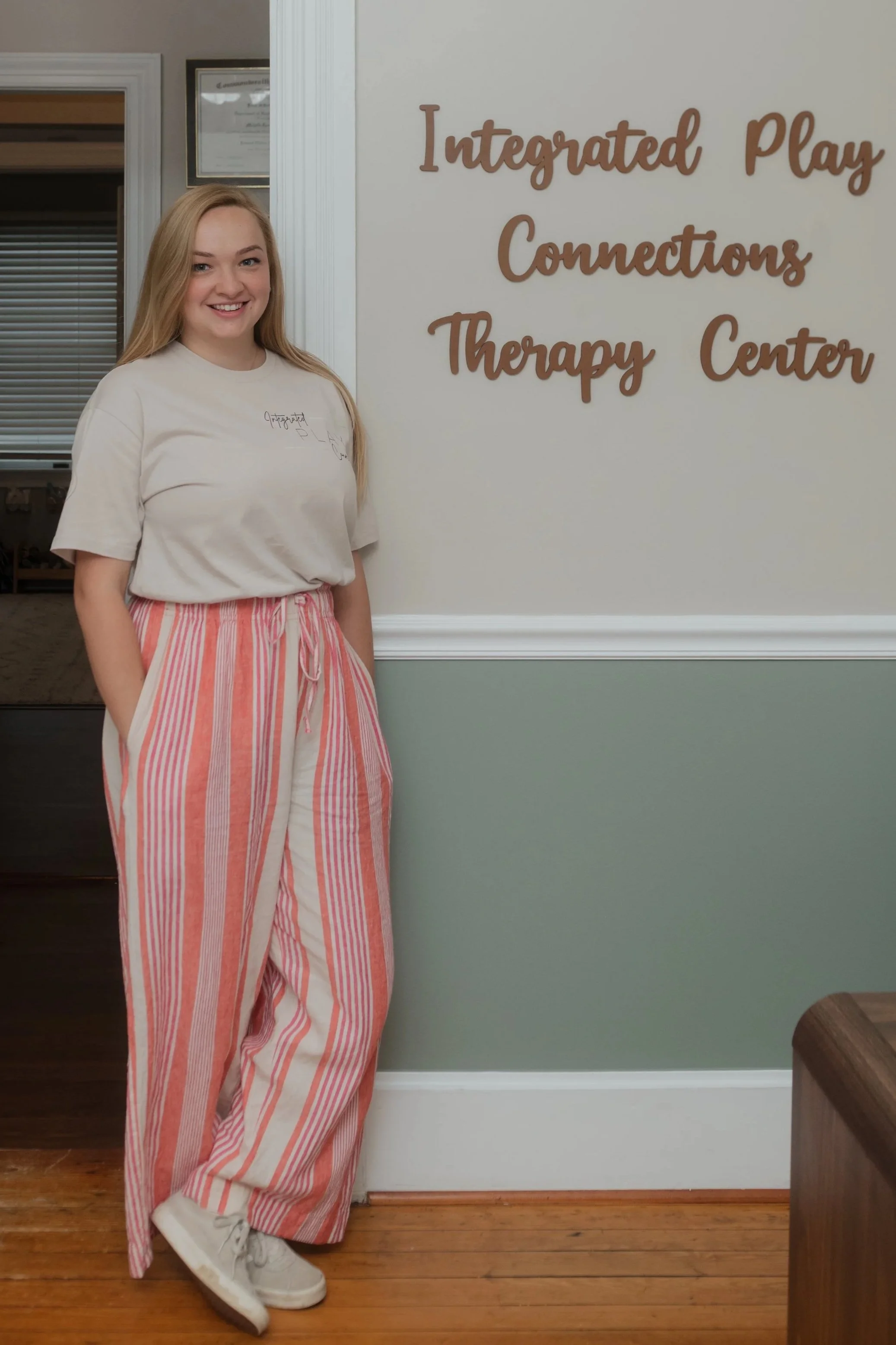 A young woman with long blonde hair smiling stands inside a therapy center, near a sign that reads 'Integrated Play Connections Therapy Center'.