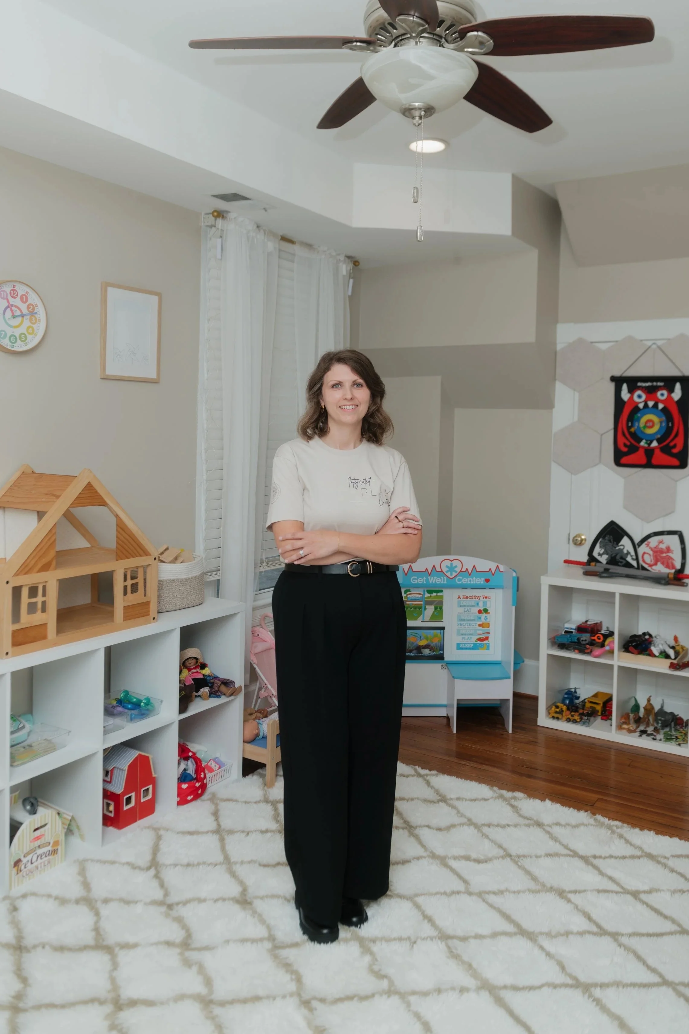 A woman standing in a child playroom, smiling, with her arms crossed. The room has white shelving filled with toys and dolls, a white and beige patterned rug, and a ceiling fan with wooden blades.