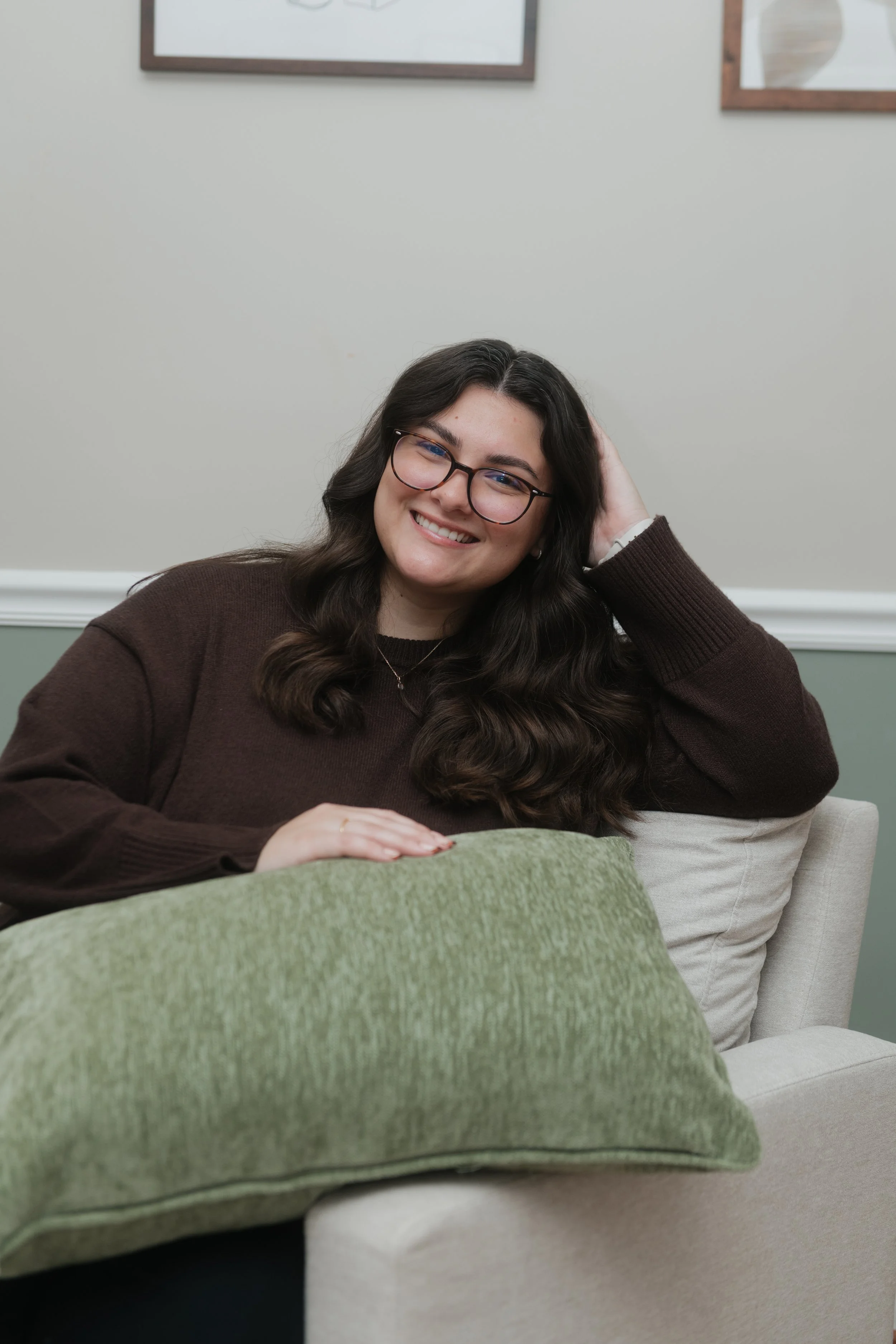 A young woman with long dark hair and glasses, smiling, seated on a beige sofa with green and beige pillows, against a light-colored wall with framed artwork.
