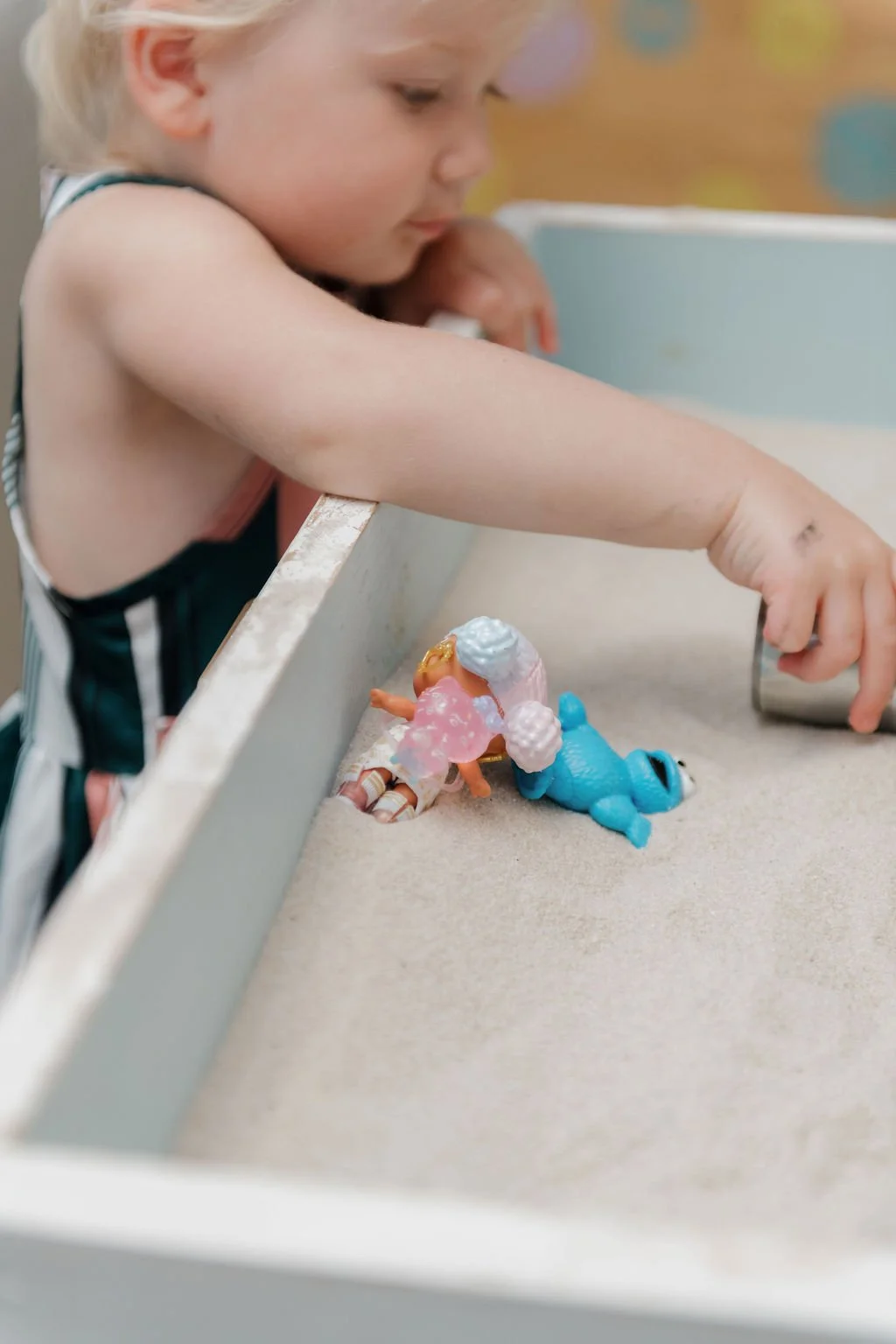 A young girl with blonde hair reaching into a box filled with small toys on a table, with a colorful background.
