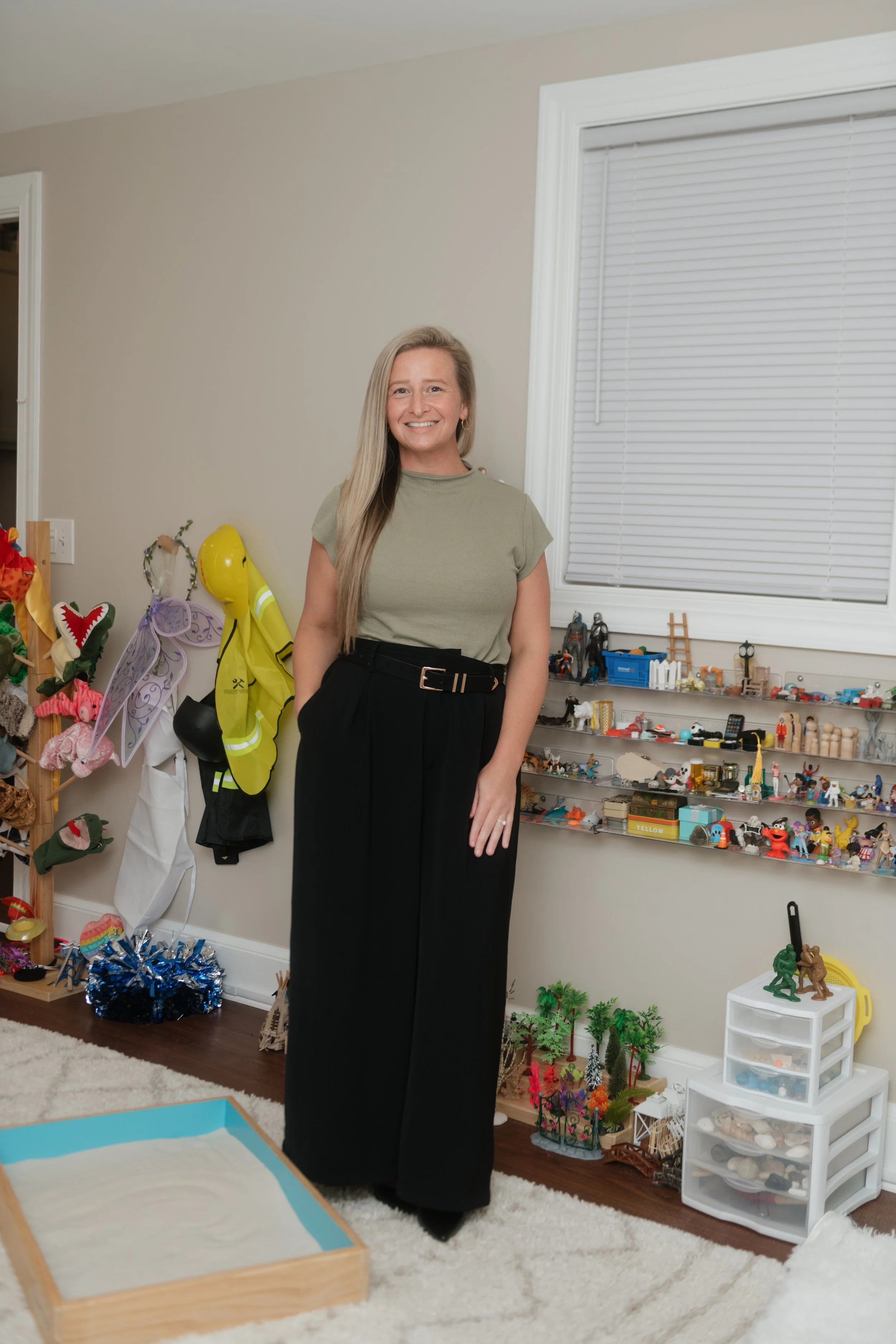 Smiling woman with long blond hair standing in a room with toys and collectibles on shelves behind her.