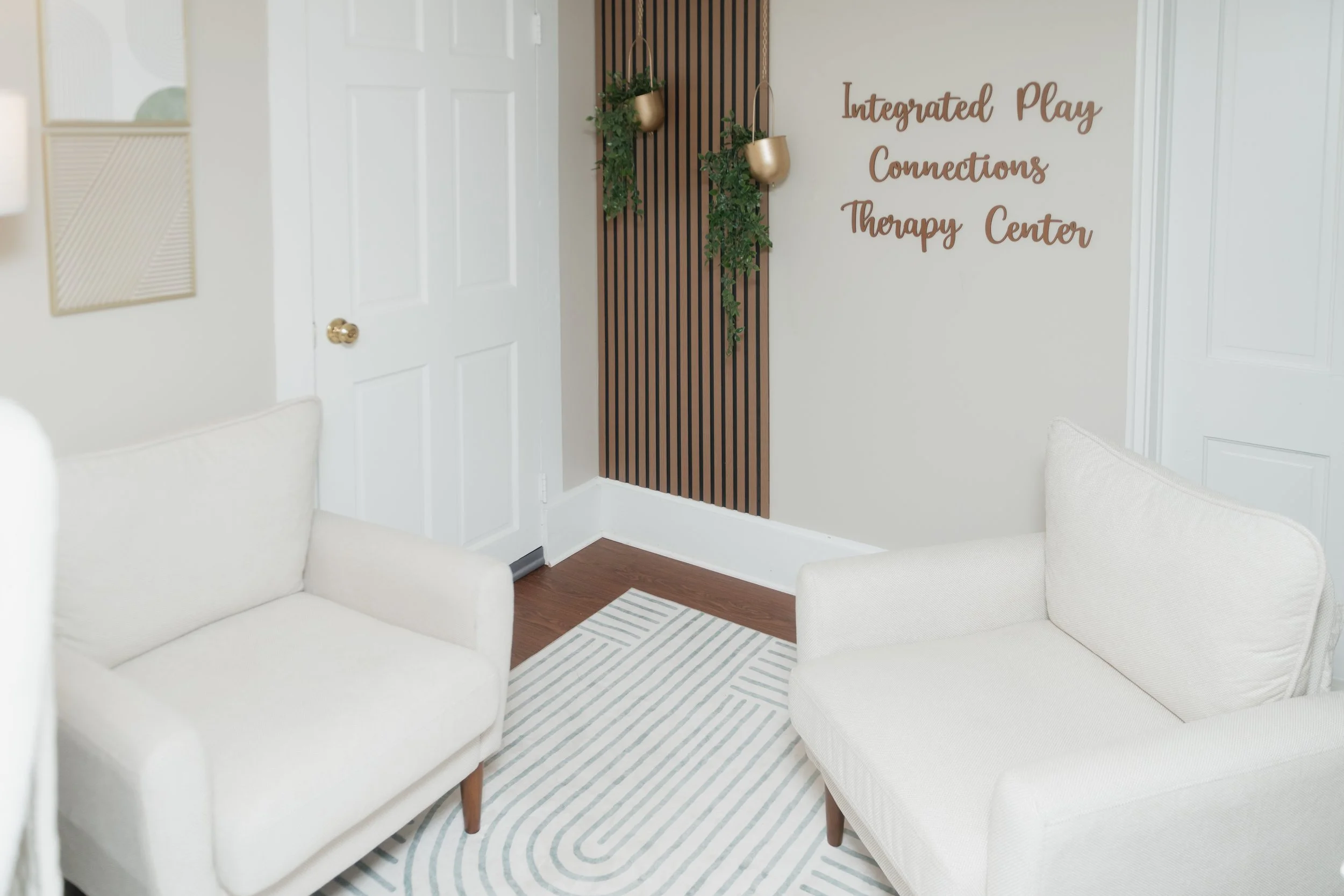 Interior of a therapy or wellness center with two white couches, a striped rug, a wooden accent wall with hanging green plants, and a sign on the wall that reads 'Integrated Play Connections Therapy Center'.