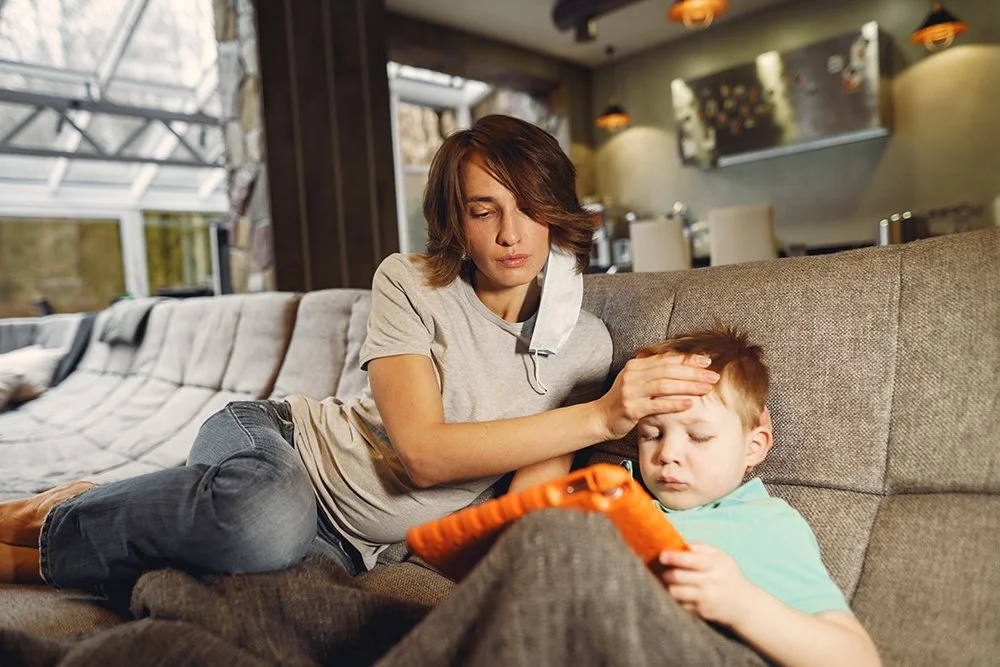 A woman is resting her hand on a young boy's forehead, who is lying on a couch and looking at a tablet, in a cozy indoor setting.