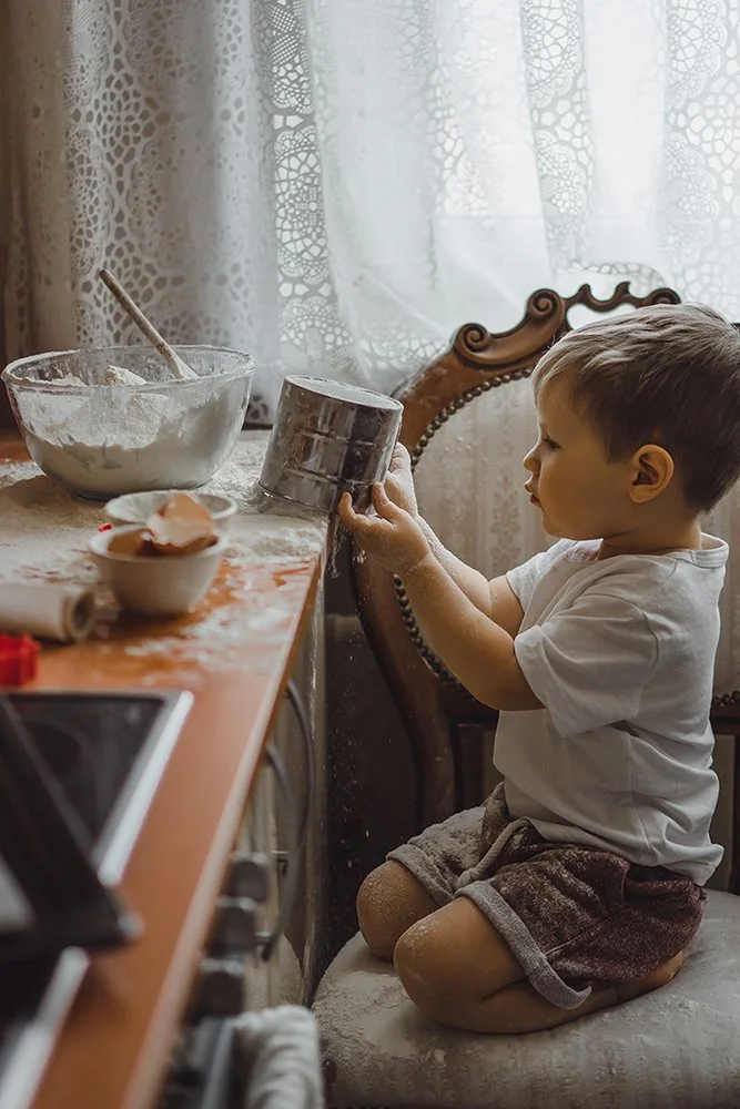 A young boy kneeling on a cushioned chair, engaging in baking or cooking. The table in front of him has a large mixing bowl, a smaller bowl with eggs, and a rolling pin. The boy is holding a metal container and appears to be pouring or mixing flour or other ingredients. The setting is a cozy kitchen with lace curtains in the background.
