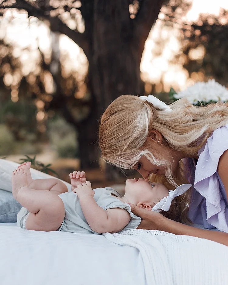 A blonde woman leaning over and touching her forehead to a baby lying on a blanket outdoors.