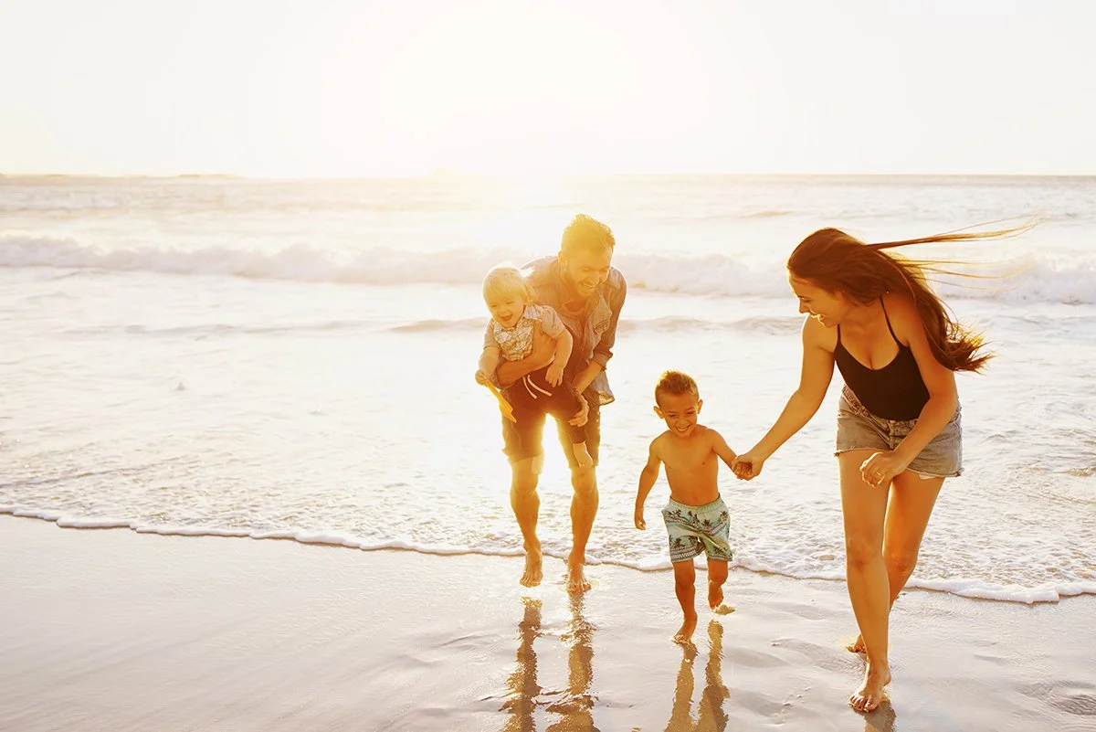 Family of four at the beach, smiling and playing near the shoreline during sunset.