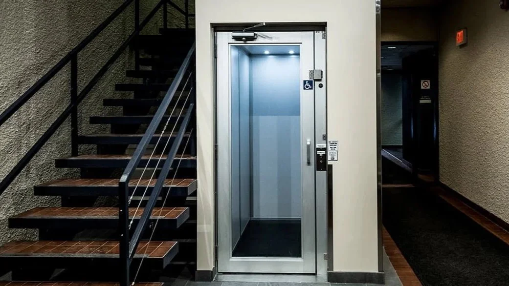 A Butler Mobility - RAM Stratus Elevator featured in a commercial building with a glass elevator door and a wood staircase to the left