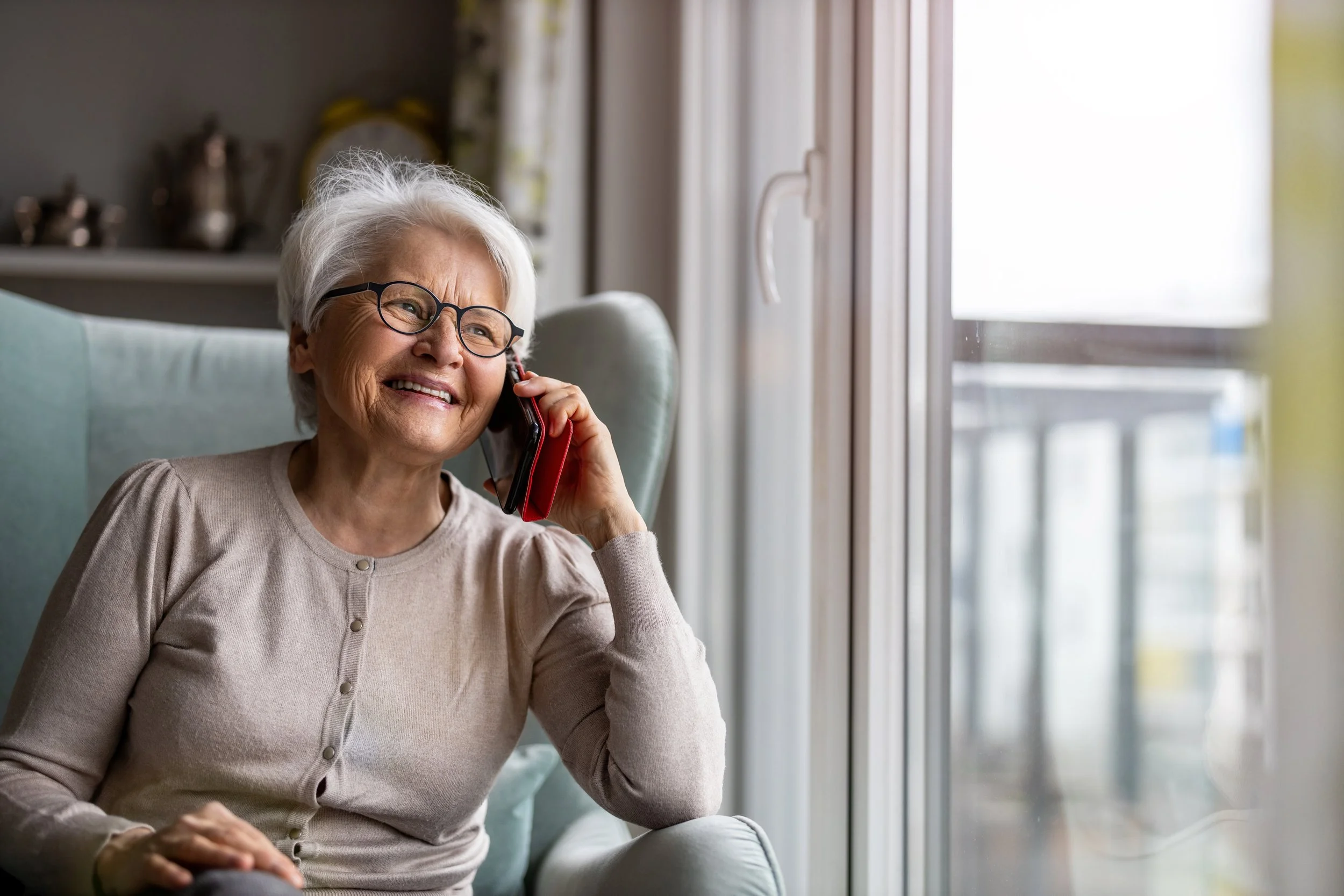 An elderly woman with white hair and glasses smiling while talking on a landline phone in a bright room near a window.