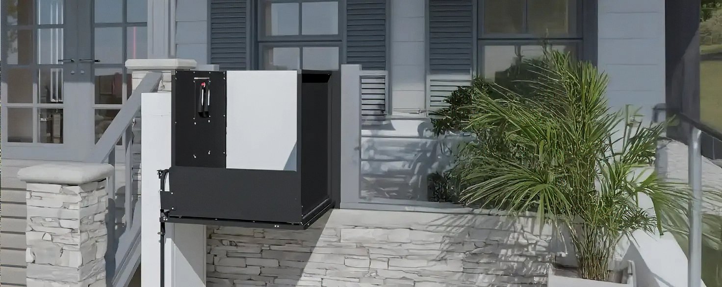A Butler Mobility - RAM vertical platform lift attached to a residential porch next to a staircase, home is white with grey shutters