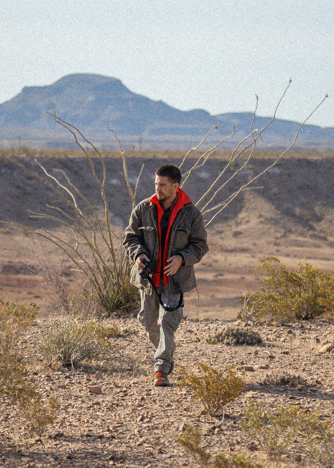 Hombre caminando en un desierto con arbustos secos y montaña al fondo.