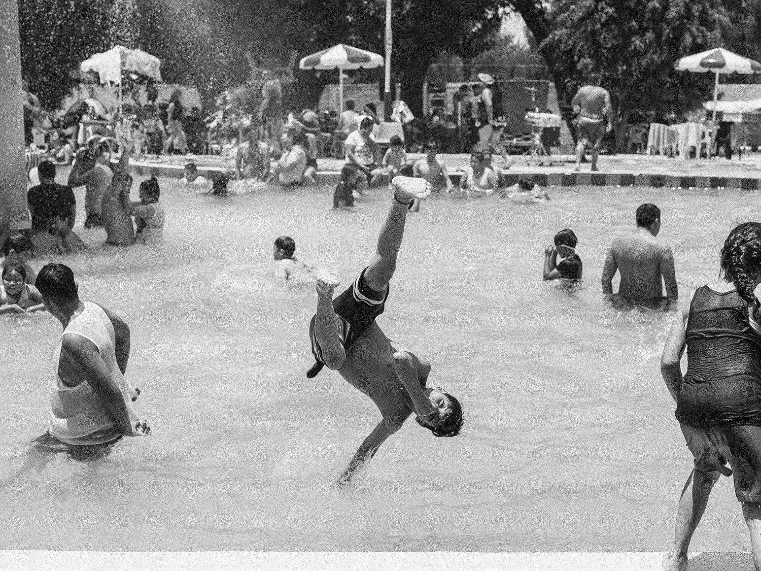 Niños y adultos disfrutando en una piscina al aire libre, con algunos niños jugando y haciendo acrobacias, mientras otros se bañan y descansan en el agua.