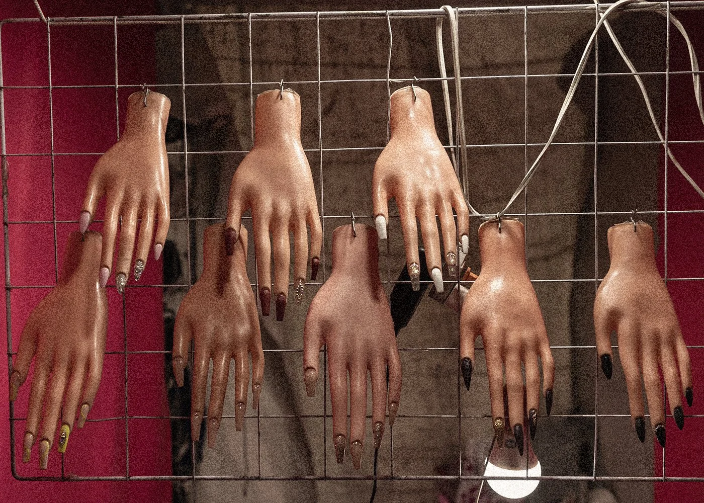 Maniquíes de manos con uñas largas pintadas en diferentes colores y diseños, colgados en una malla metálica.