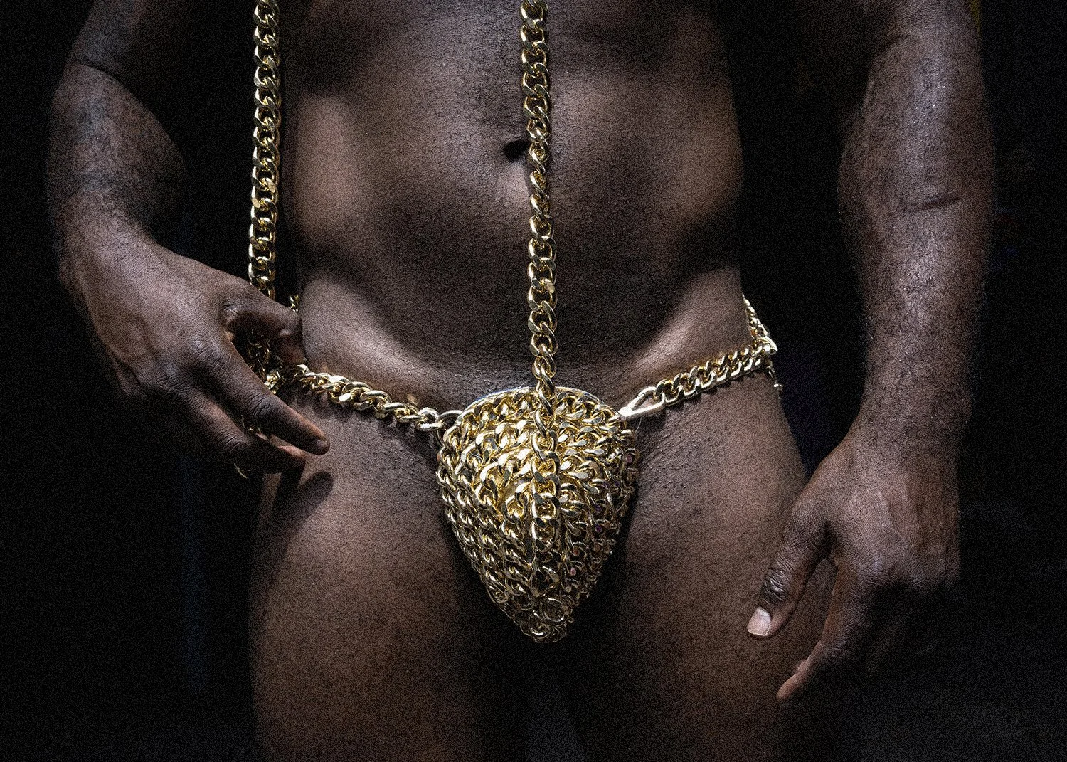 Hombre negro con piel desnuda usando múltiples cadenas de oro alrededor de la cintura y colgando en el abdomen, con las manos en las caderas.