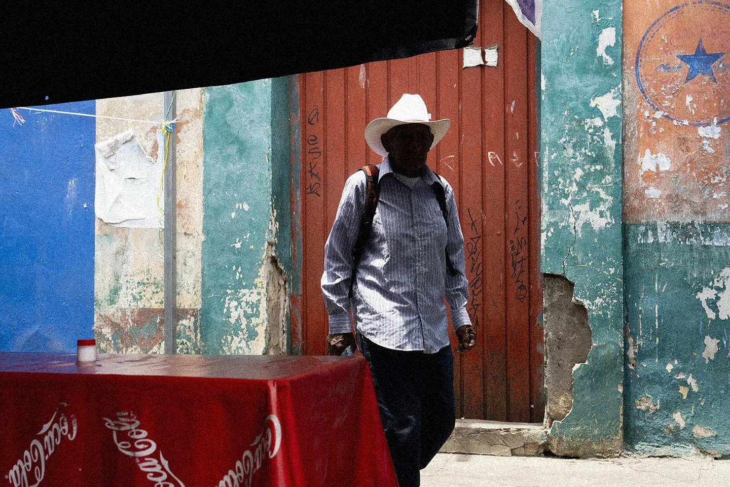 Hombre con sombrero blanco, camisa a rayas, pantalones oscuros y mochila, caminando por una calle con paredes desgastadas y graffiti, al lado de una mesa con manta roja con el logo de Coca-Cola