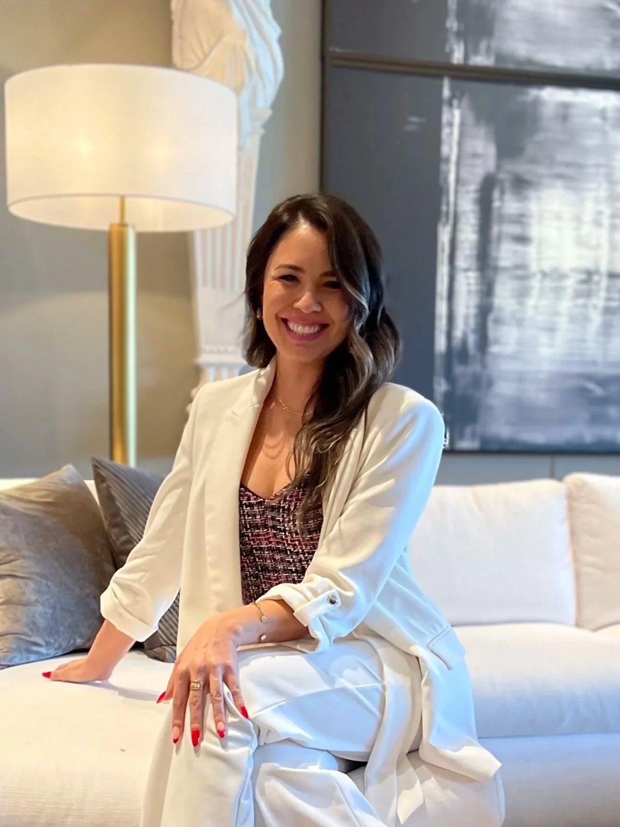 Bilingual speech language pathologist, owner of LINGUABLOOM PLLC. A woman with dark brown, wavy hair, smiling and sitting on a cream-colored sofa in a well-lit living room. There is a modern lamp and abstract artwork in the background.