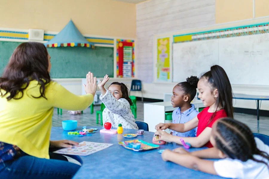 A teacher and four young students sitting at a table in a classroom. The teacher is giving a high-five to one student who is smiling. The classroom has colorful decorations, a whiteboard, and educational materials.