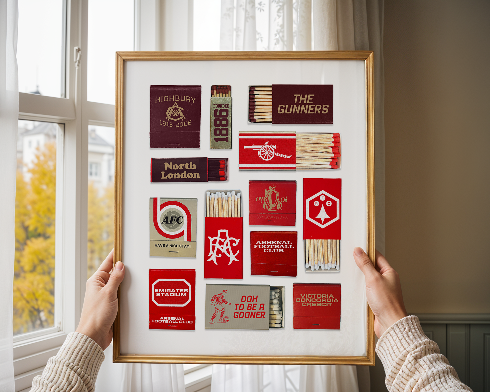 A person holding a framed display of matchboxes decorated with soccer club logos such as Highbury, The Gunners, North London, Arsenal Football Club, and others, with a window showing an autumn scene outside.