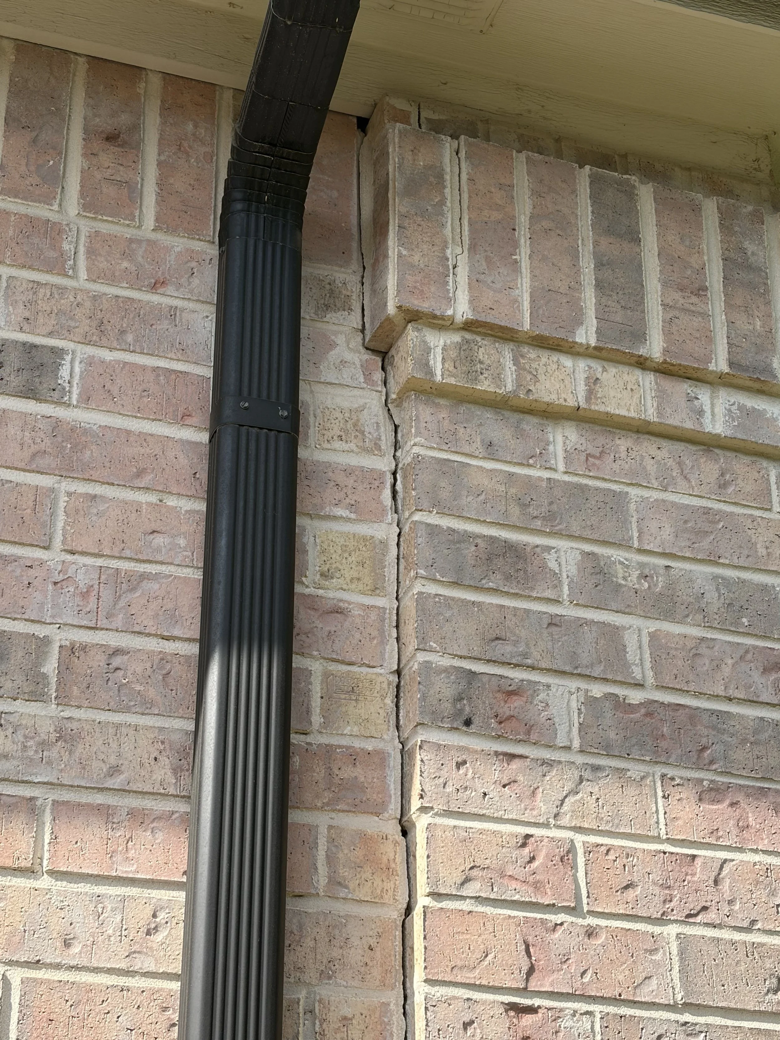 Close-up of a brick exterior wall with a vertical crack at the corner intersection, and a black downspout pipe running along the wall.