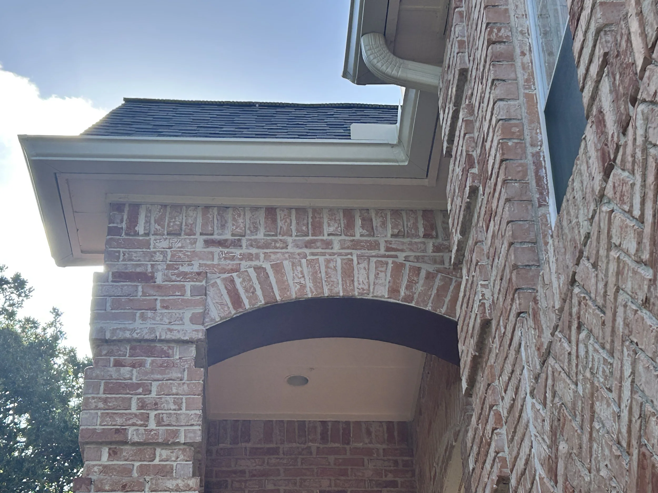 Close-up image of a brick building with an arched doorway and a window, with clear blue sky and some trees visible in the background.