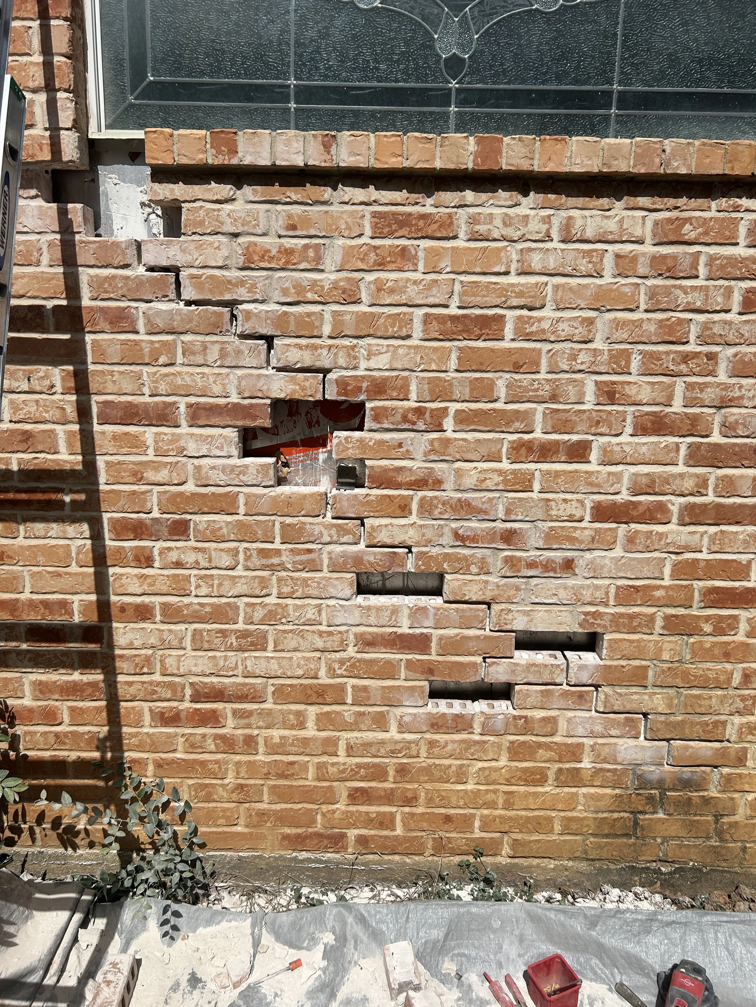 Part of a brick wall under construction with some bricks removed, exposing gaps, and construction tools and materials on the ground in front.