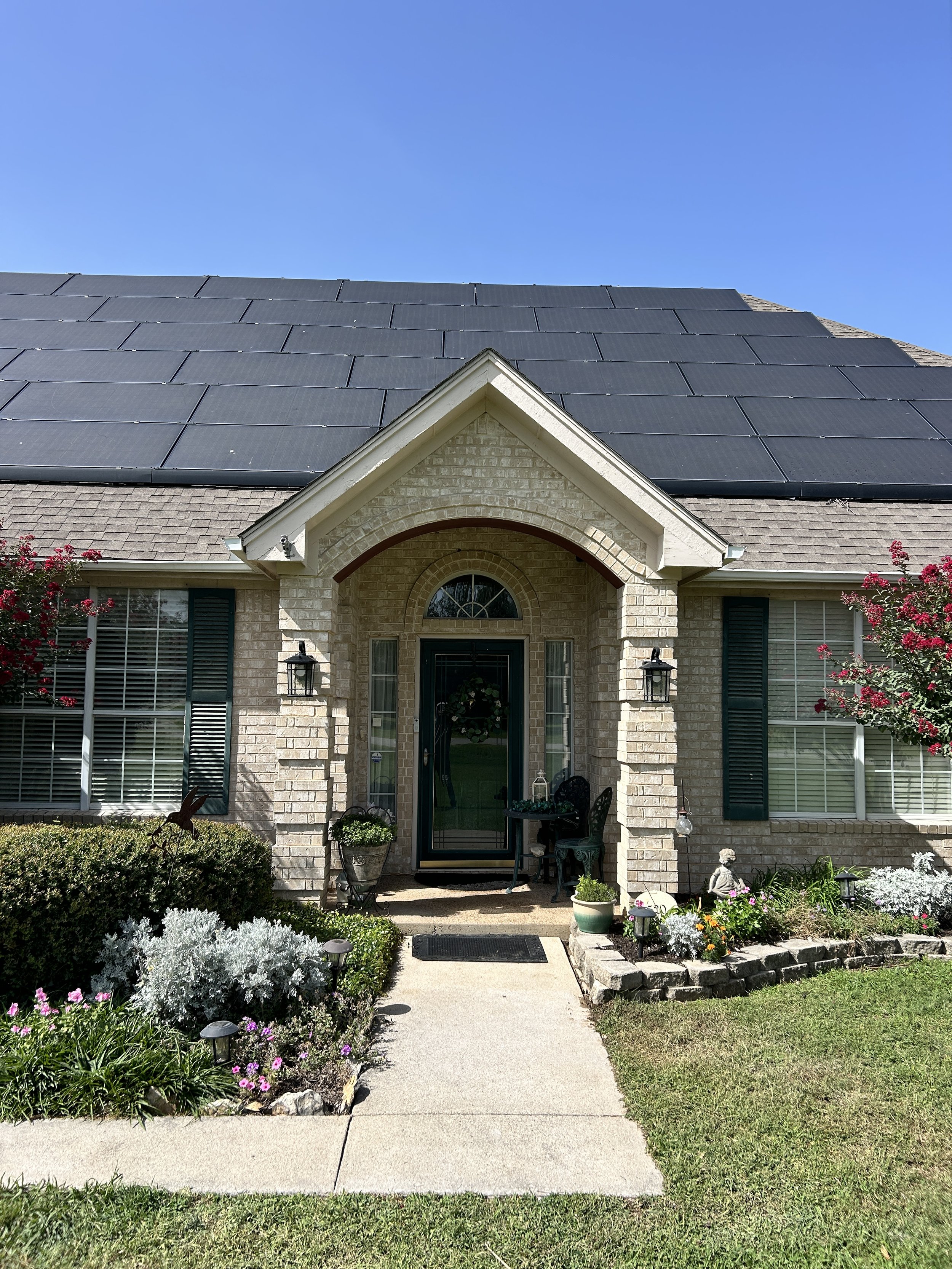 Front view of a brick house with a peaked roof covered in solar panels, a porch with a door and window, a garden with flowers, shrubs, and garden ornaments, and a sidewalk leading to the entrance under a clear blue sky.