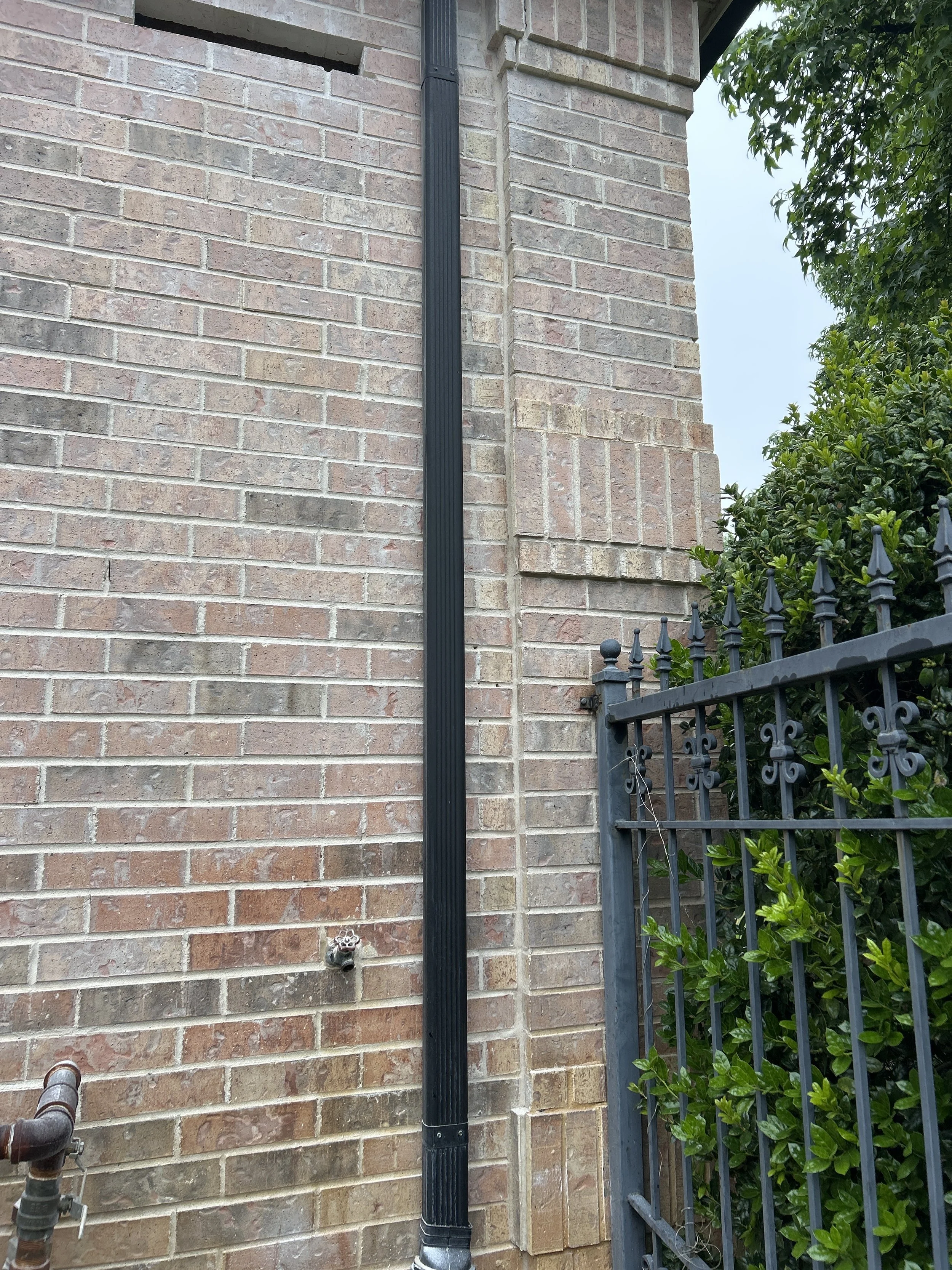 A black downspout attached to a brick wall, next to a black metal fence with decorative elements, and some green shrubs on the right side of the image.