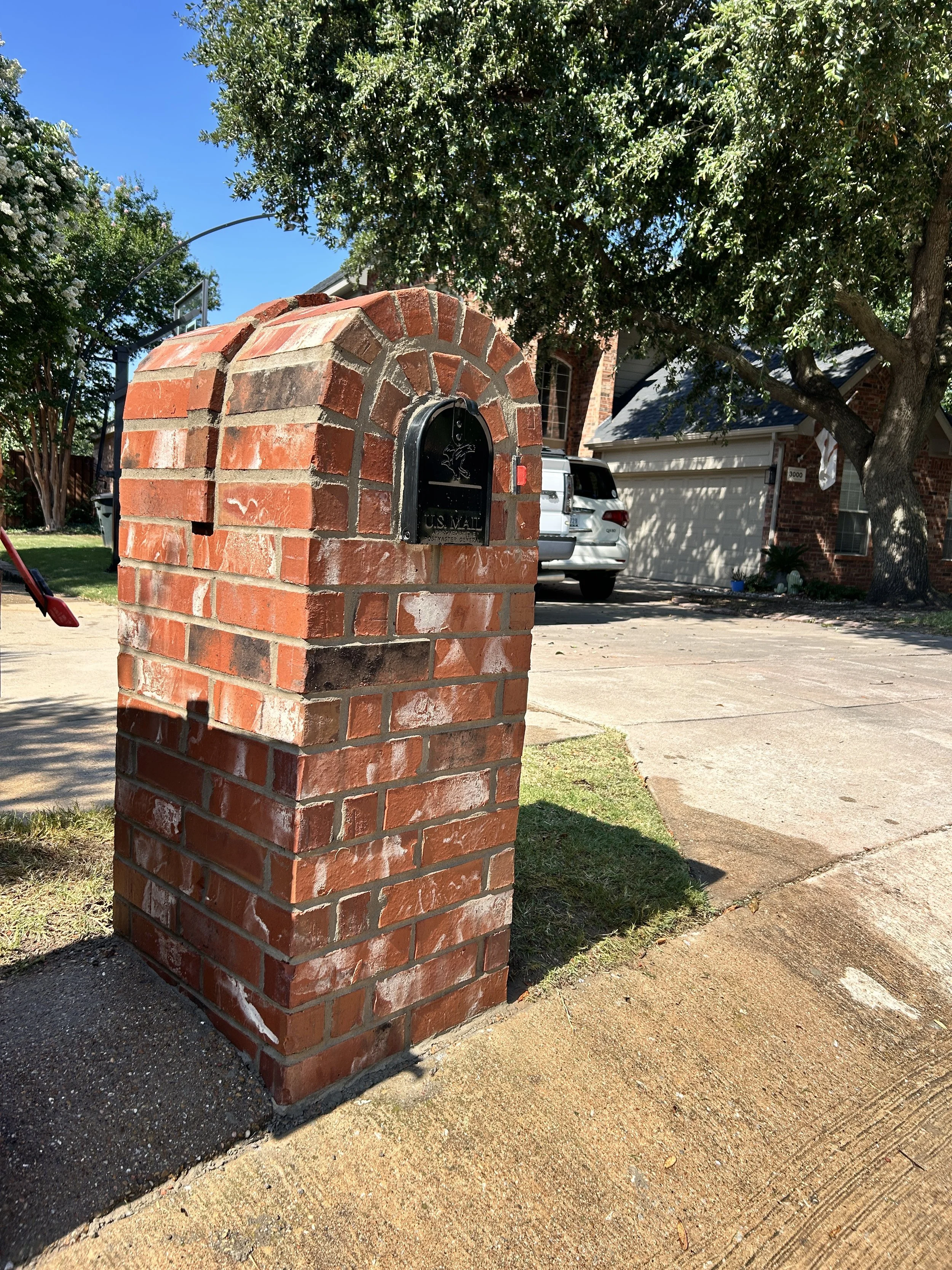 Close-up of a red brick mailbox with a black door, in a suburban neighborhood, with trees and a house in the background.