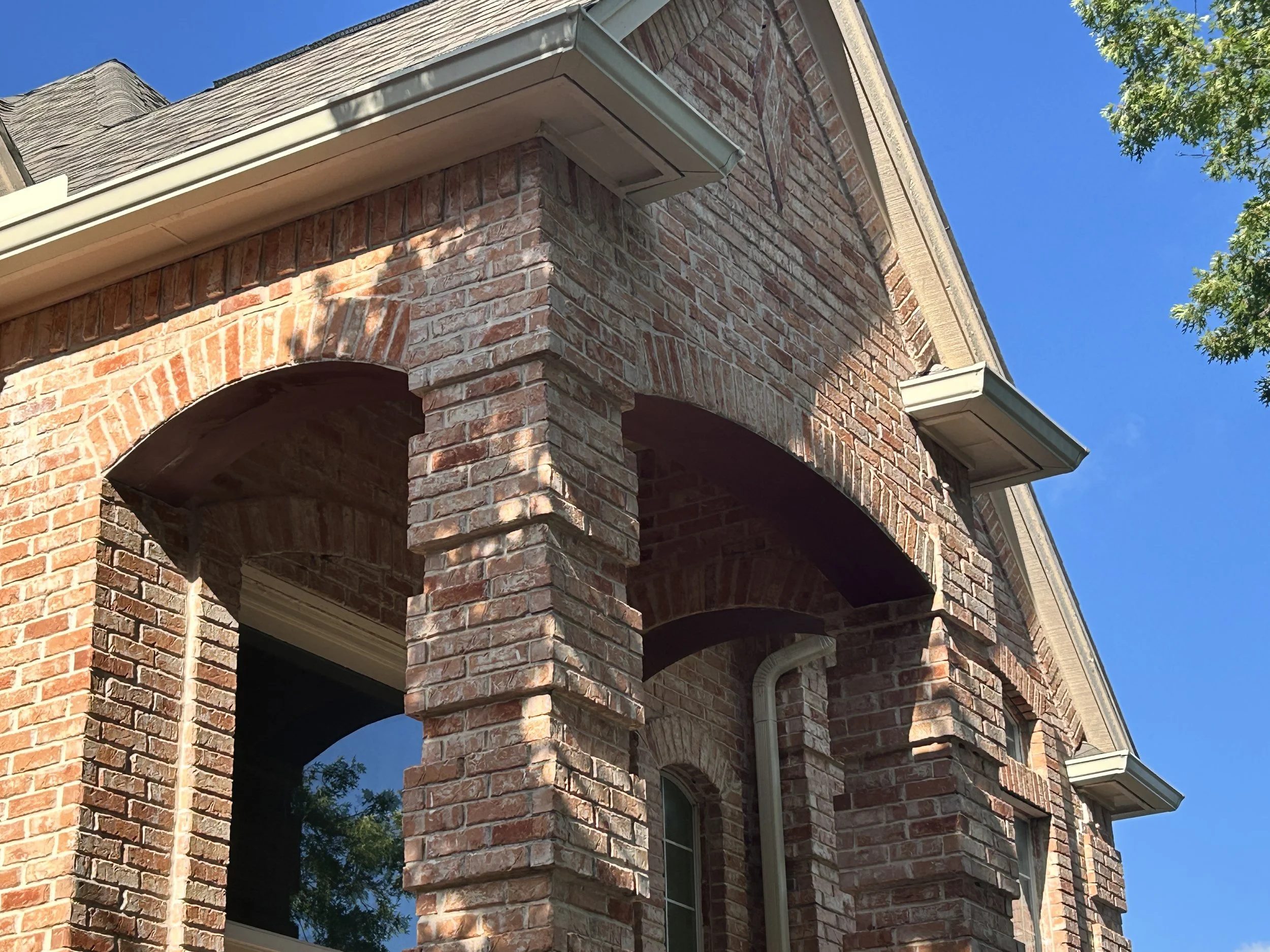 Close-up of a brick house's corner with arched openings, beige trims, and a blue sky background.