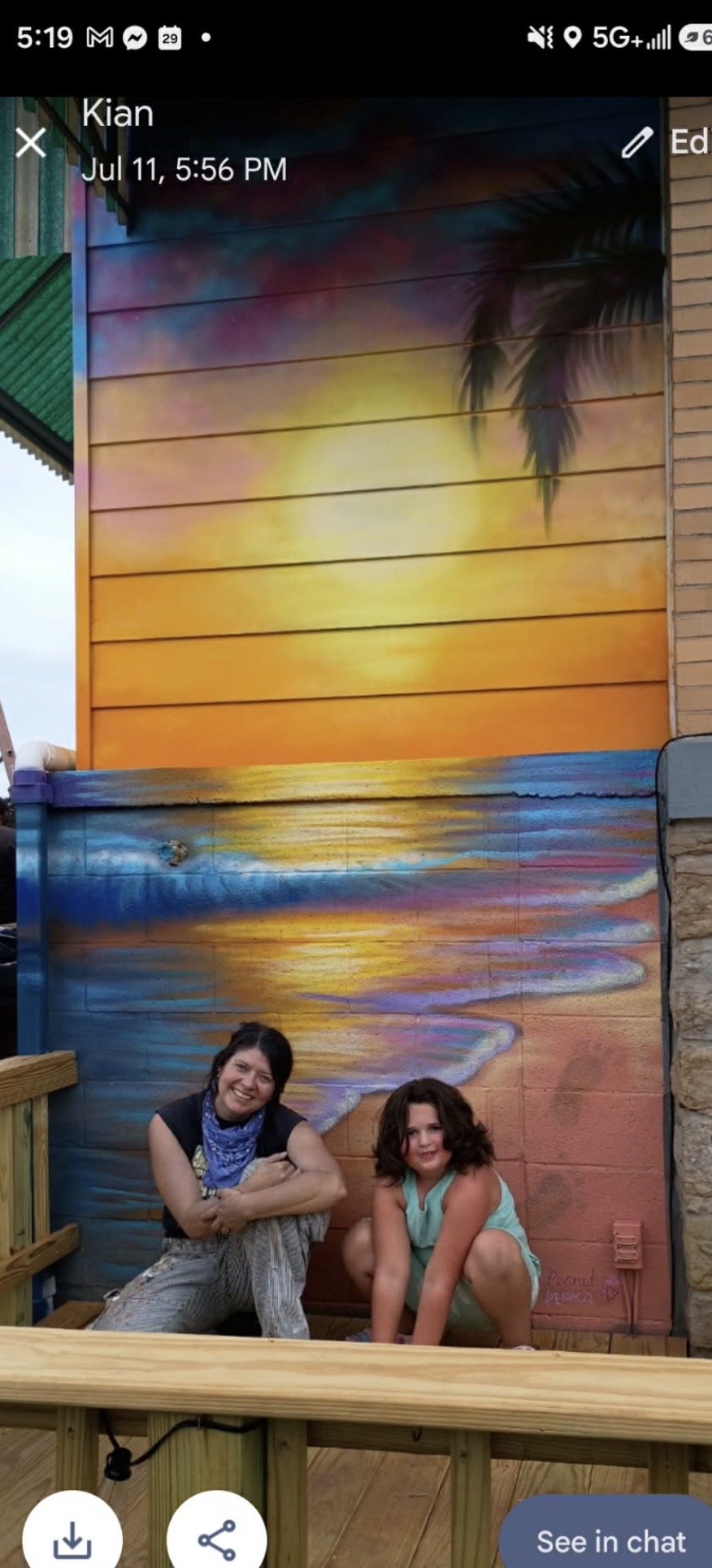 A woman and a girl sitting in front of a colorful sunset mural with a palm tree silhouette, on a wooden deck.