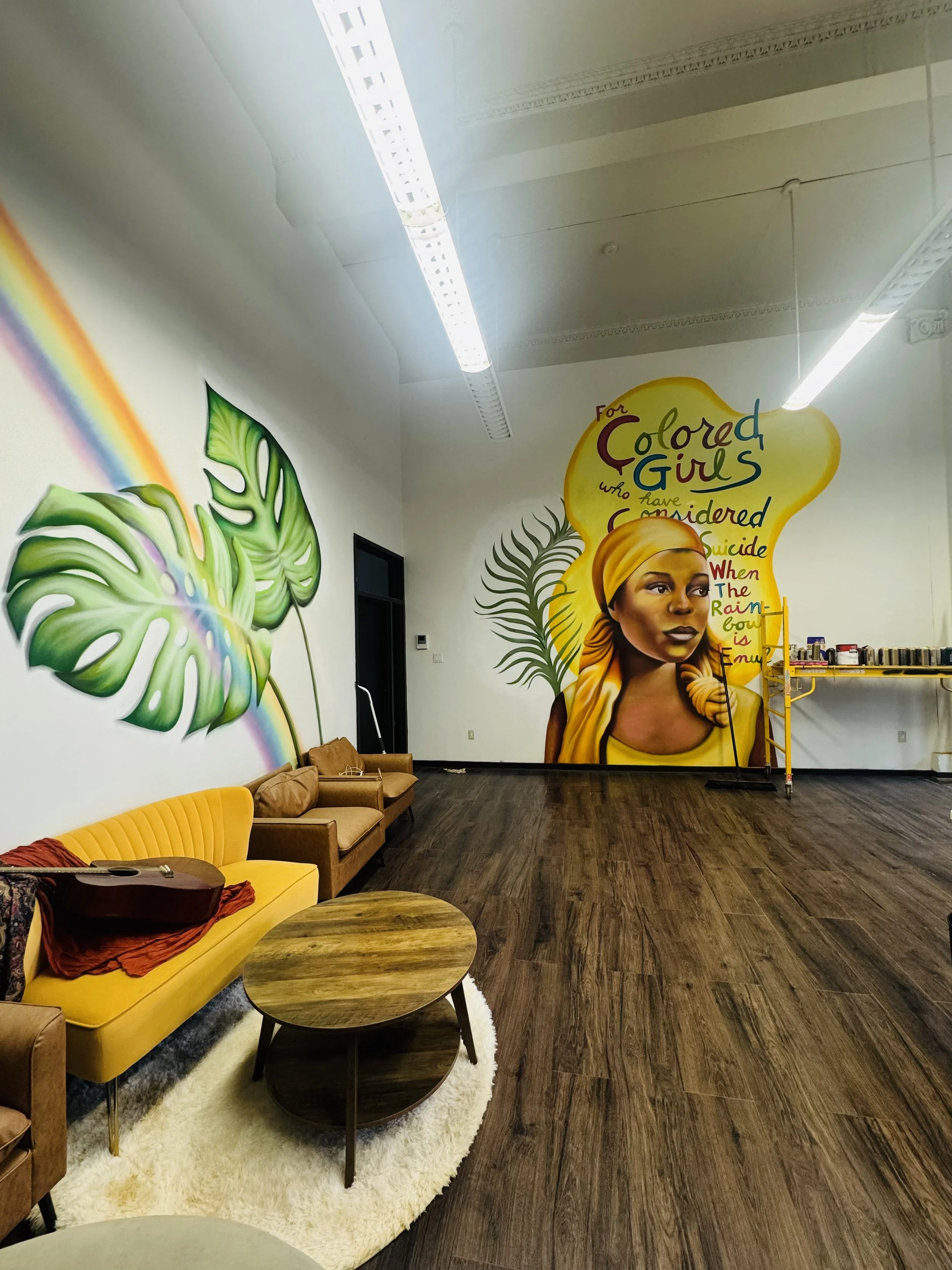 Interior view of a room with mural art, colorful furniture including a yellow couch, a round wooden table, a cream-colored armchair, and a white shag rug. The mural features large green leaves, a woman with a yellow headscarf, and vibrant text.