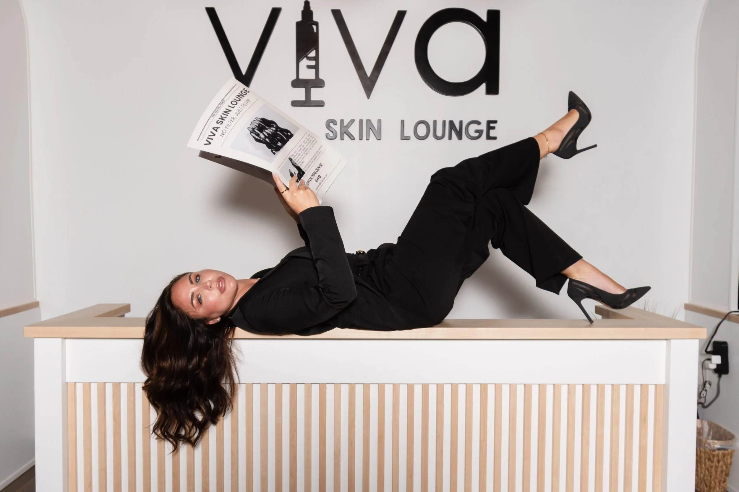 Woman wearing black outfit and high heels lying on a desk with one leg crossed over the other, reading a newspaper, set against a wall with black letters spelling 'VIVA SKIN LOUNGE' and a syringe icon.