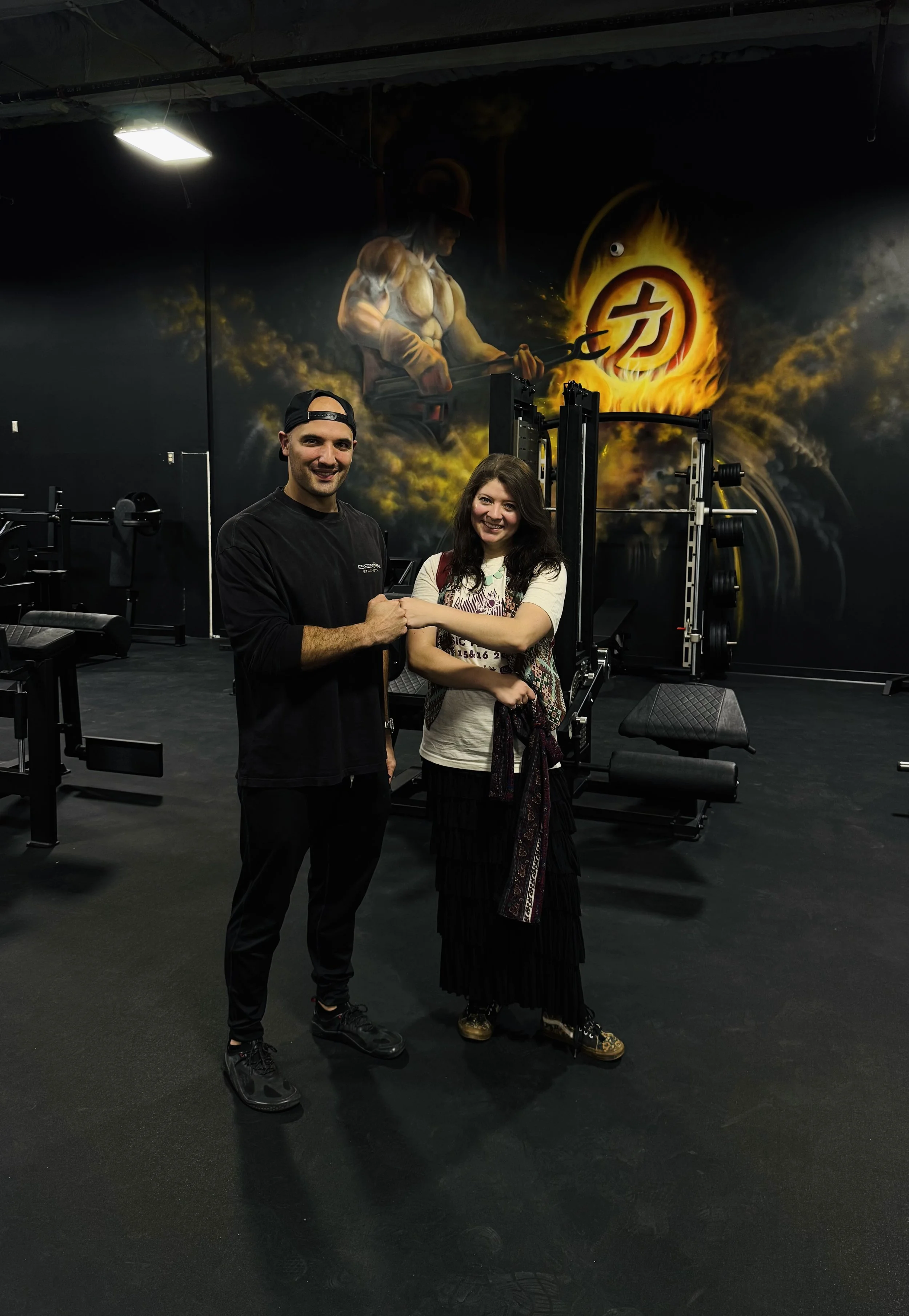 Two people shaking hands in a gym with fitness equipment and a large mural of a muscular figure holding a wrench against a fiery background.