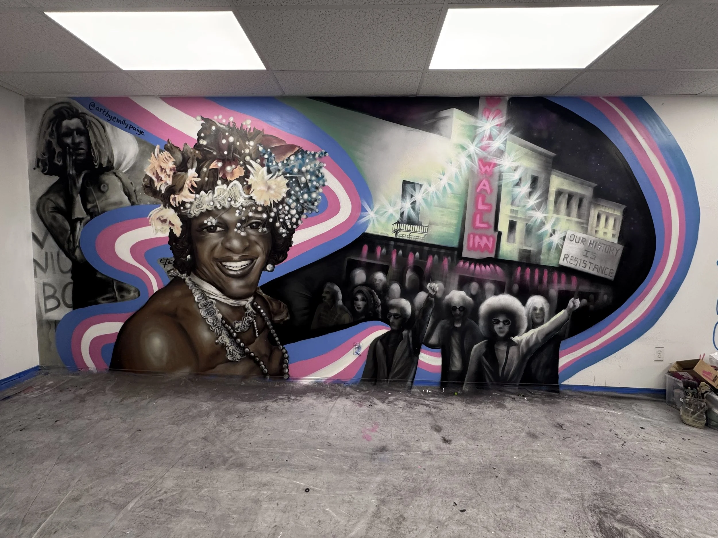 Colorful mural on a wall depicting a smiling woman with a beaded necklace and floral headpiece, alongside various figures and buildings, with a neon pink sign reading 'DOLL IN' and other graffiti.