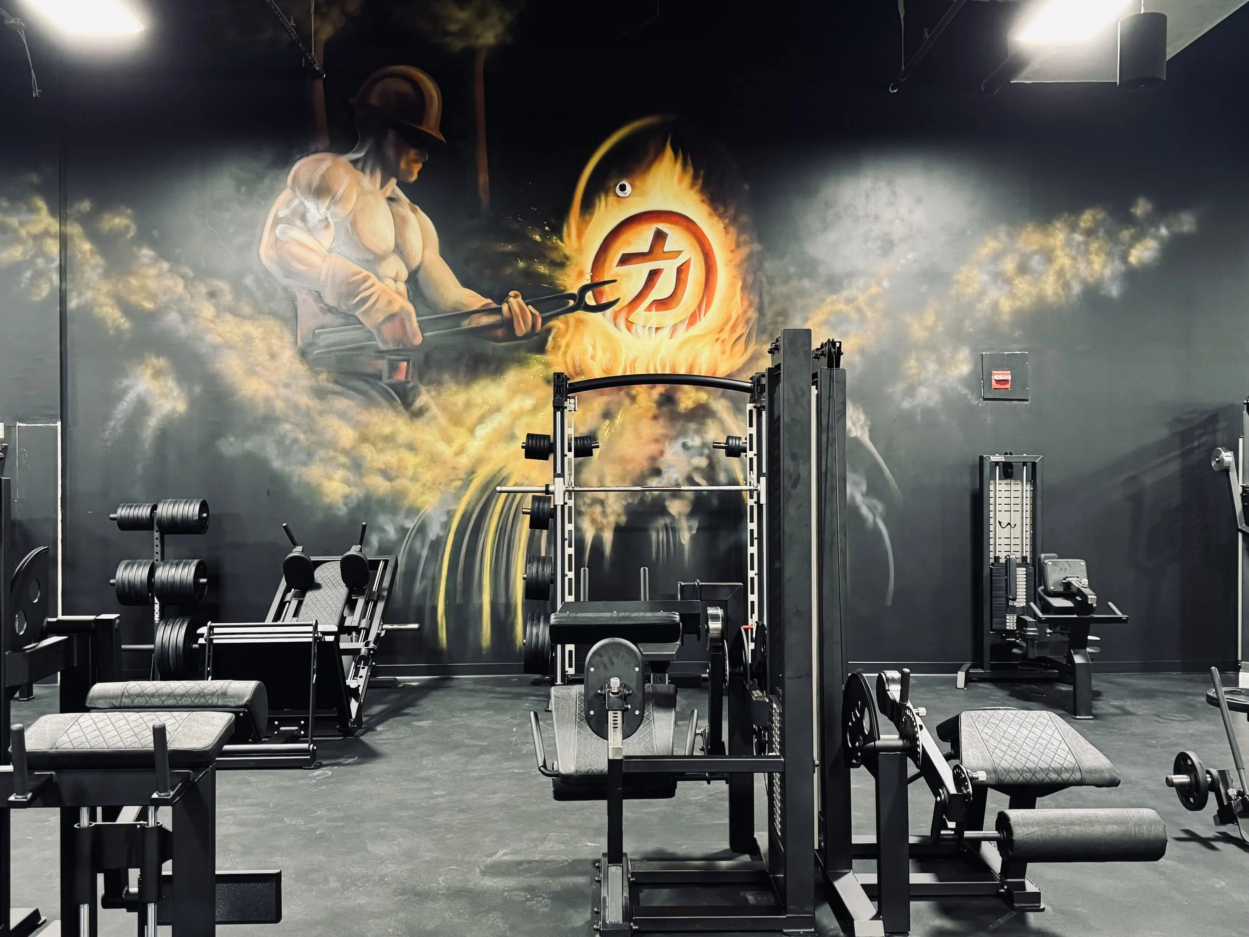 Gym with a large wall mural of a muscular soldier in a helmet holding a trident, surrounded by flames and a fiery logo, gym equipment such as weights and benches in the foreground.